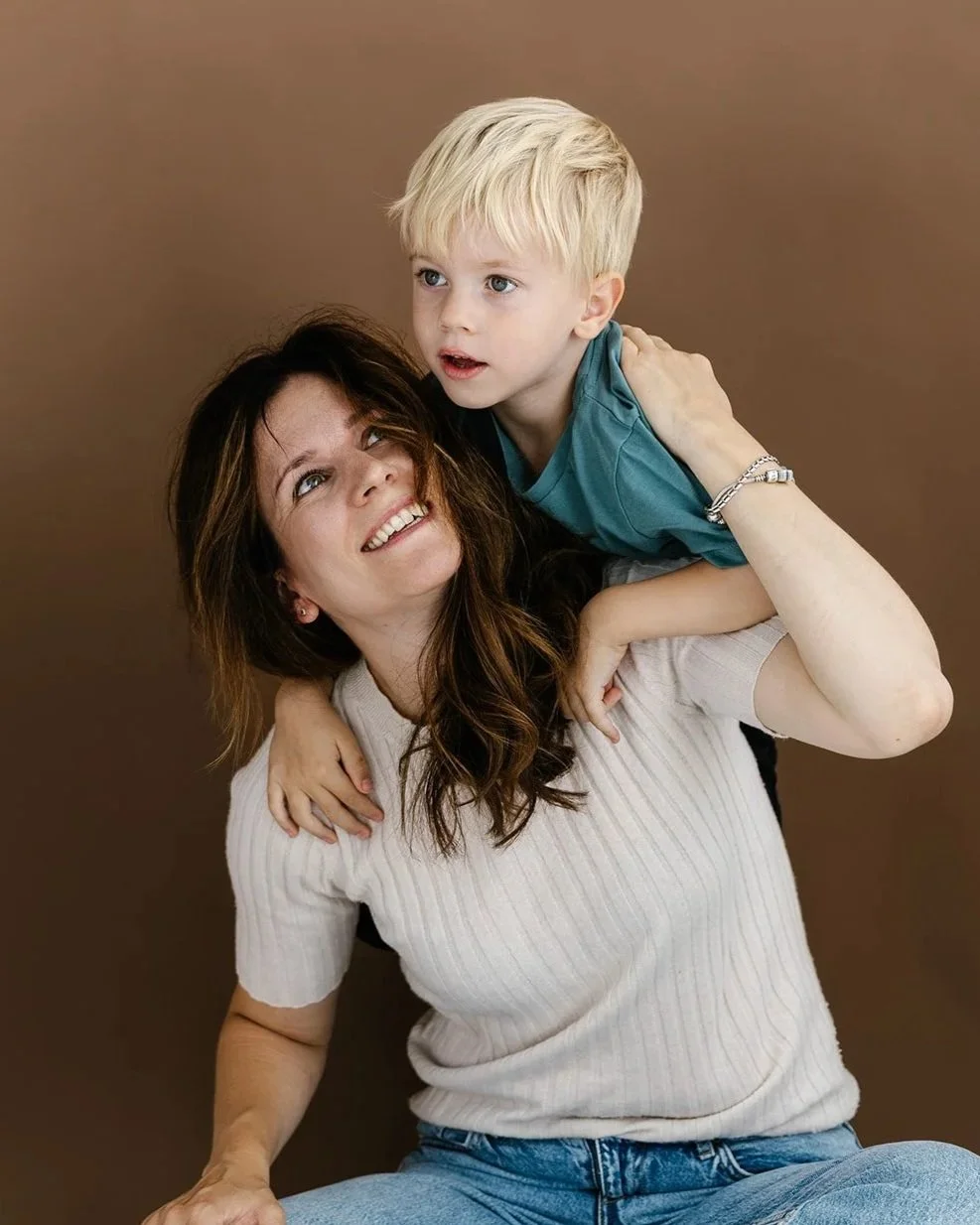 Mamma sitter på golvet med sin son på axlarna och tittar leende upp på honom.