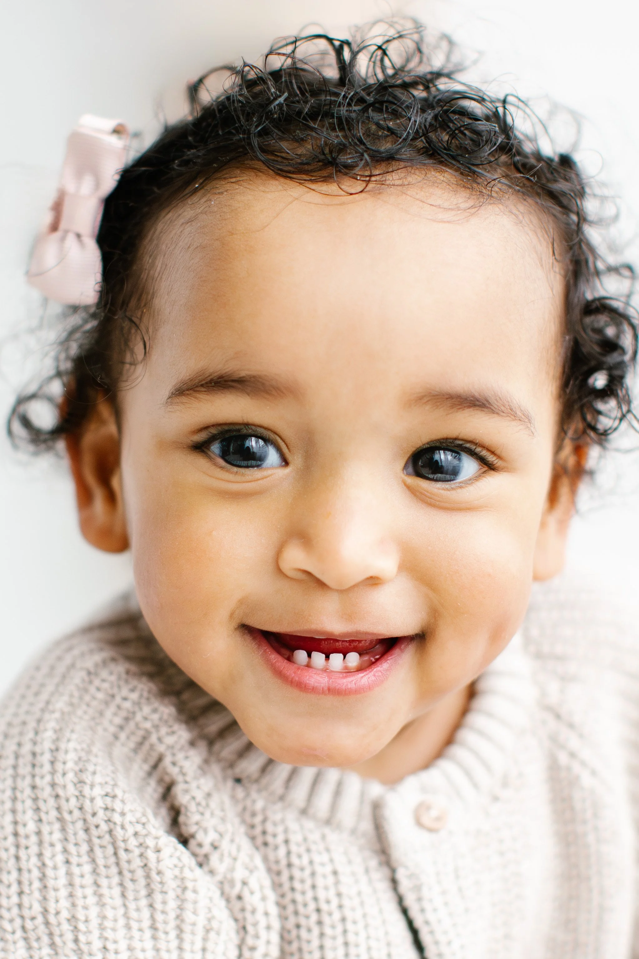 En glad liten tjej med brunt lockigt hår som har en rosett i håret.