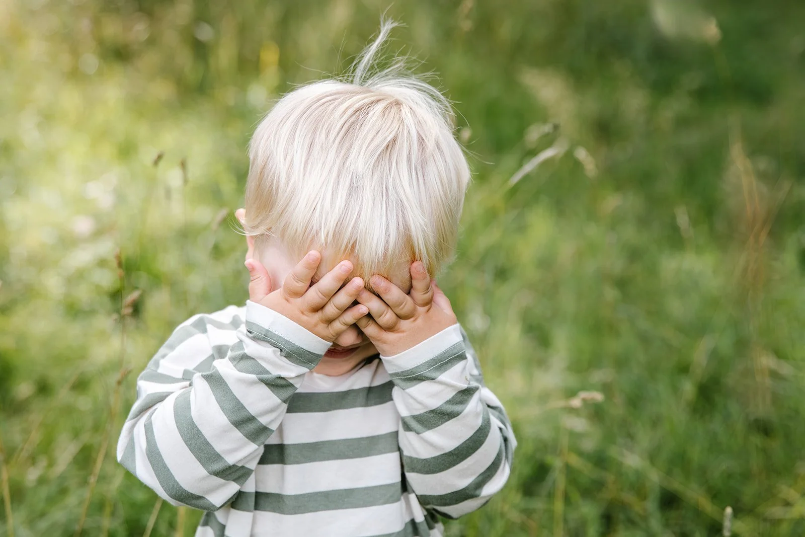 blond pojke står i högt gräs och håller för ögonen. han har randig tröja