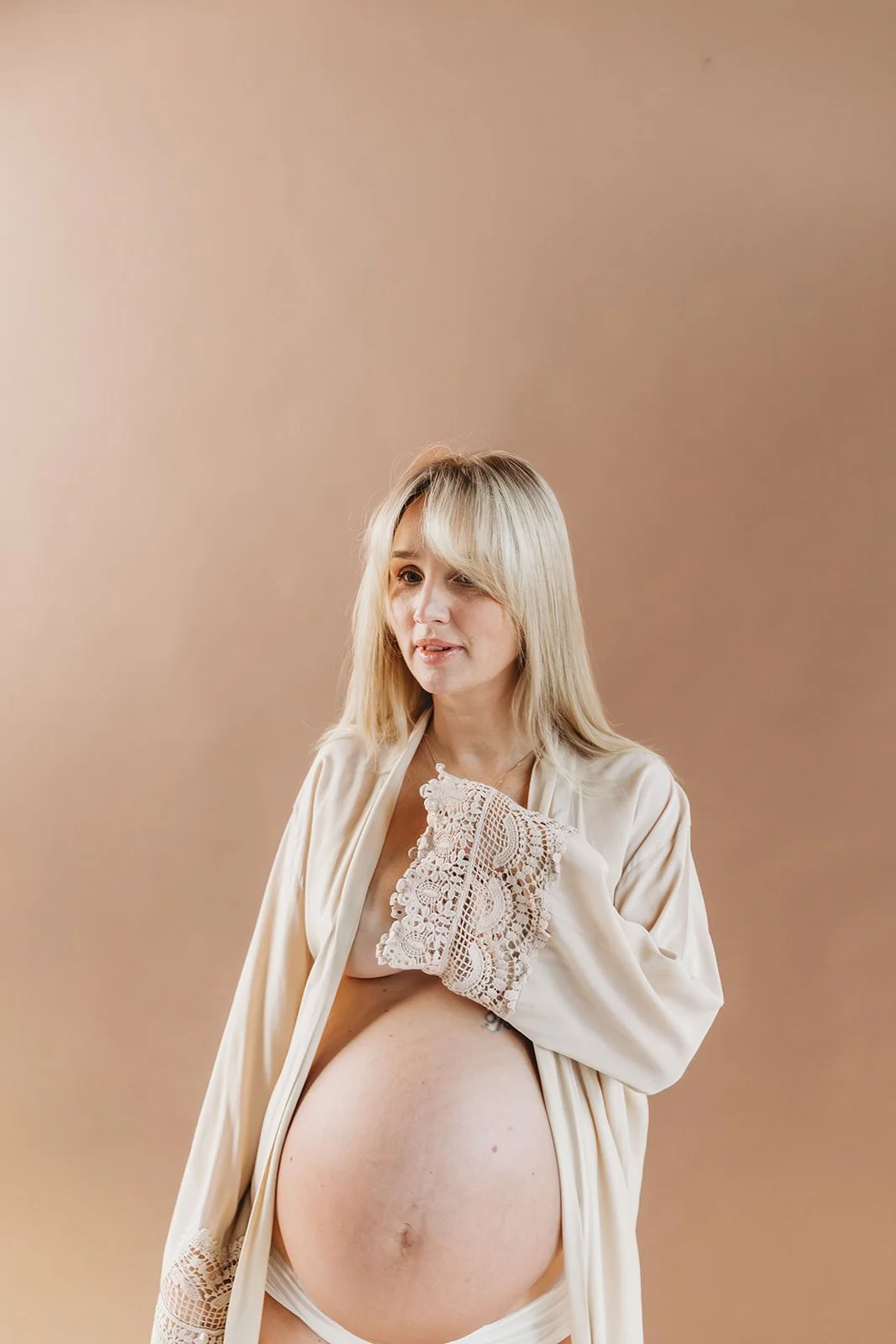 En gravid kvinna med långt blont hår, som bär en beige kimono, står mot en beige bakgrund.