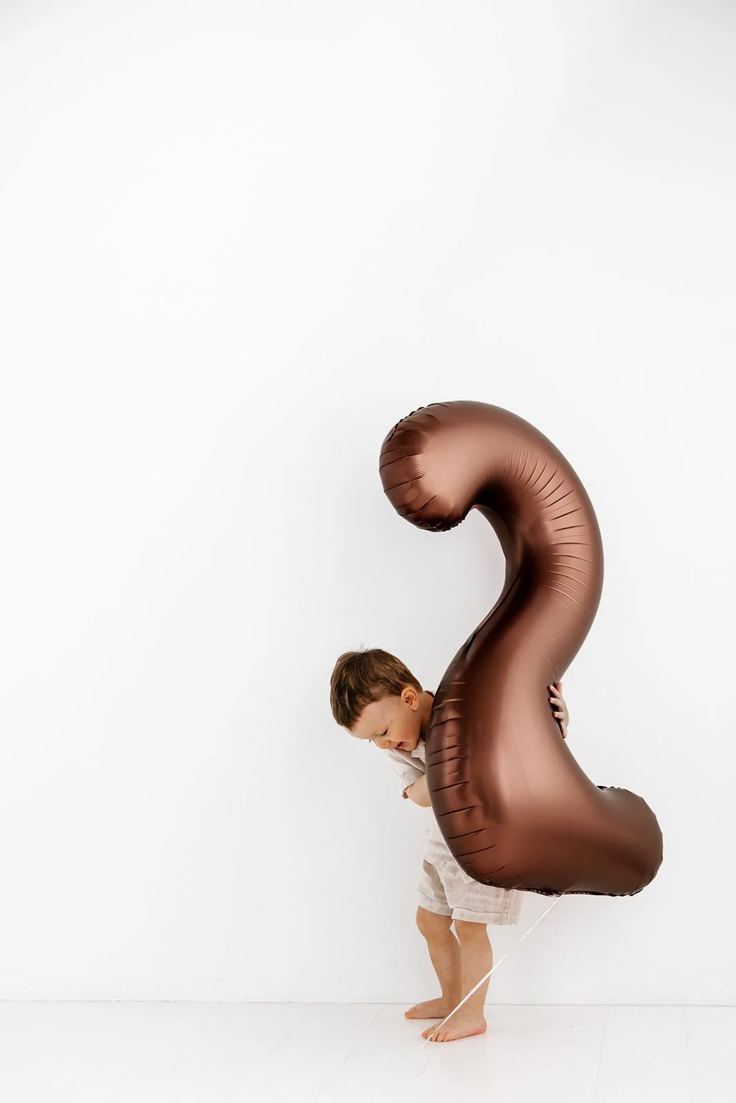 En ung pojke håller en stor heliumballong i form av siffran två. Han är klädd i ljusa shorts och en vit tröja, och står mot en vit bakgrund.