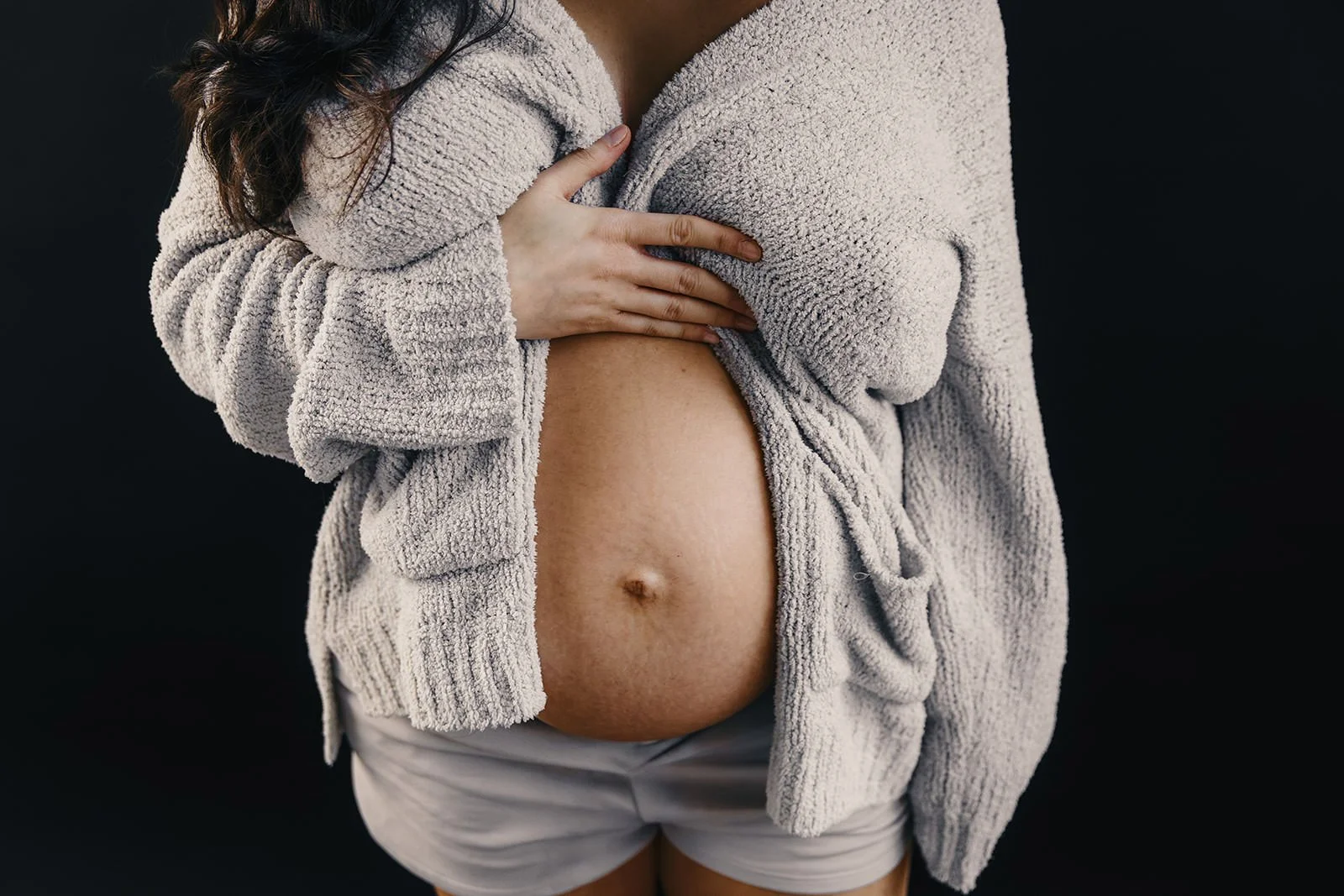 En kvinna som bär en beige, fluffig tröja och vita shorts, håller handen på sin gravidmage, fotad mot en svart bakgrund.