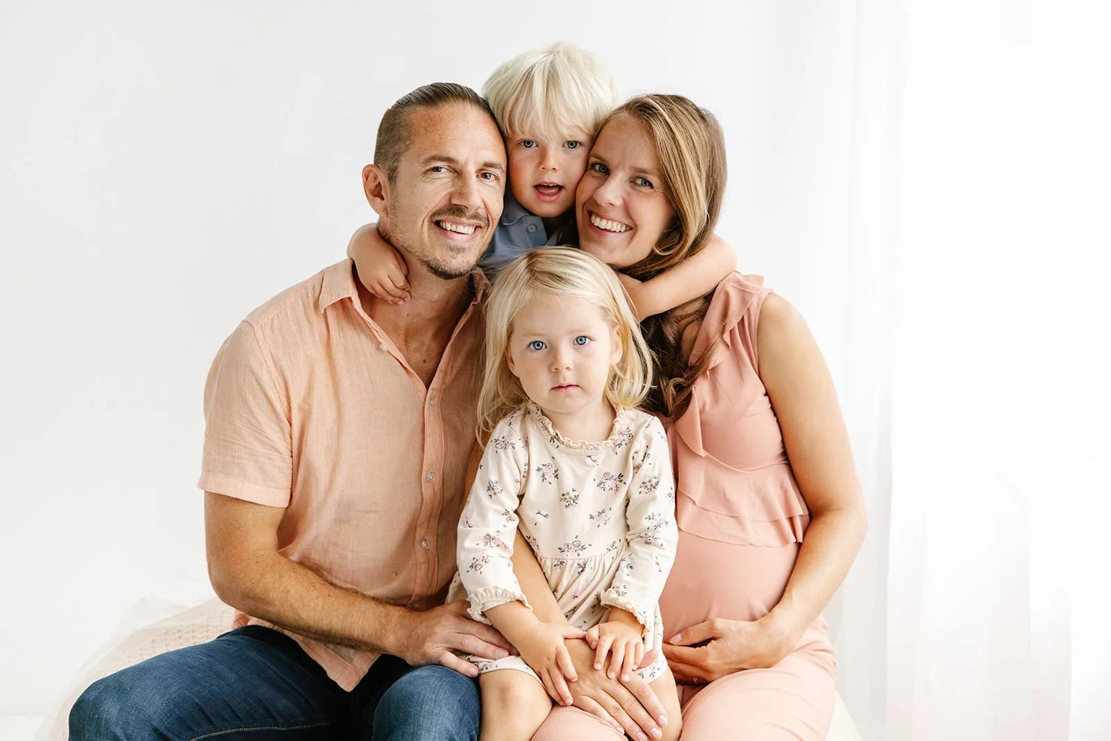 familj med mamma, pappa, dotter och son i förskoleåldern kramar om varandra under en fotografering i studio. Bakgrunden är vit.