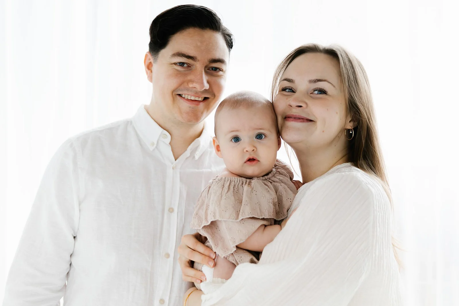 familj med dotter tittar in i kameran och ler. de har ljusa kläder och bakgrunden är vit.