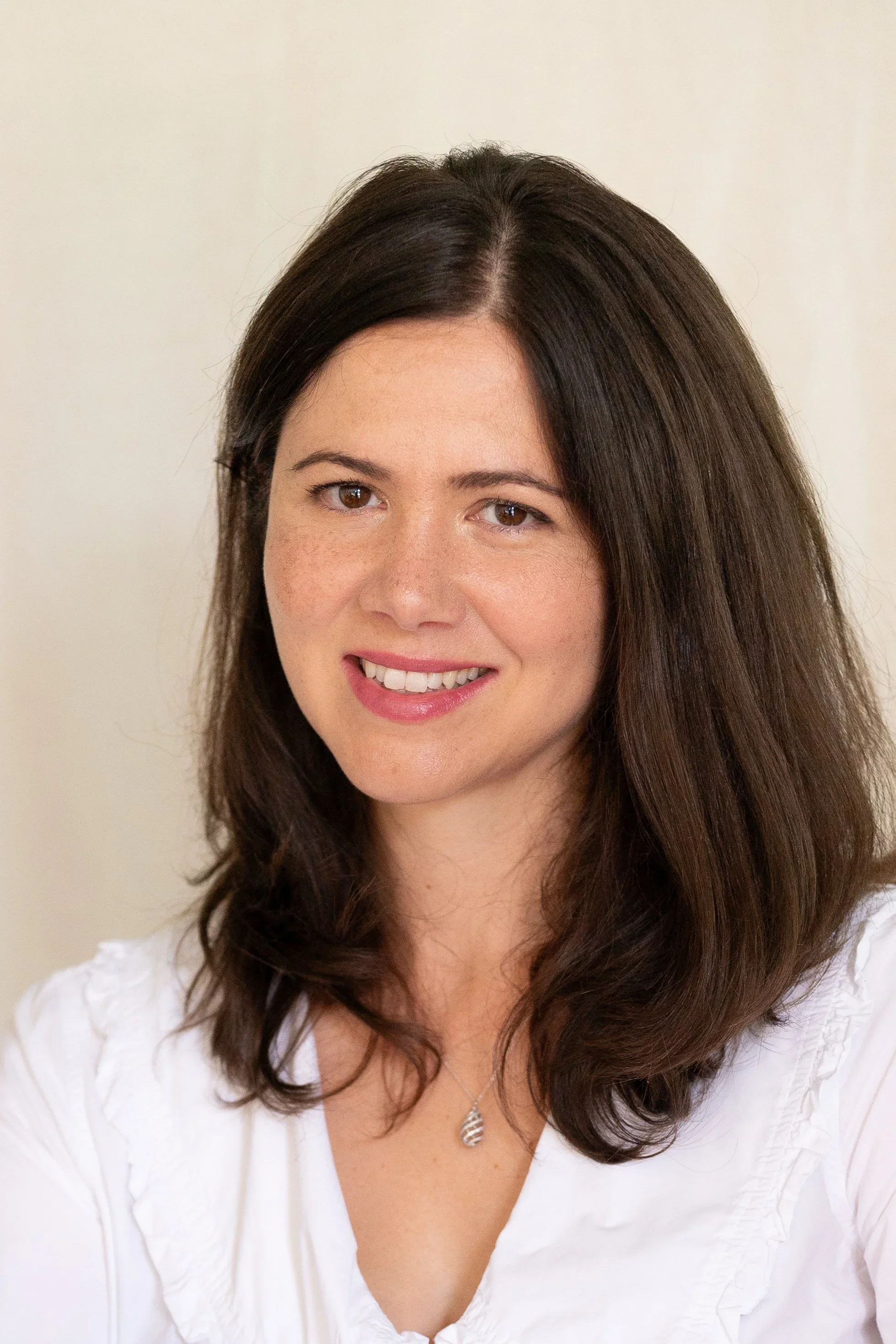A woman with shoulder-length dark brown hair, wearing a white blouse and a silver necklace, smiling at the camera against a plain light-colored background.