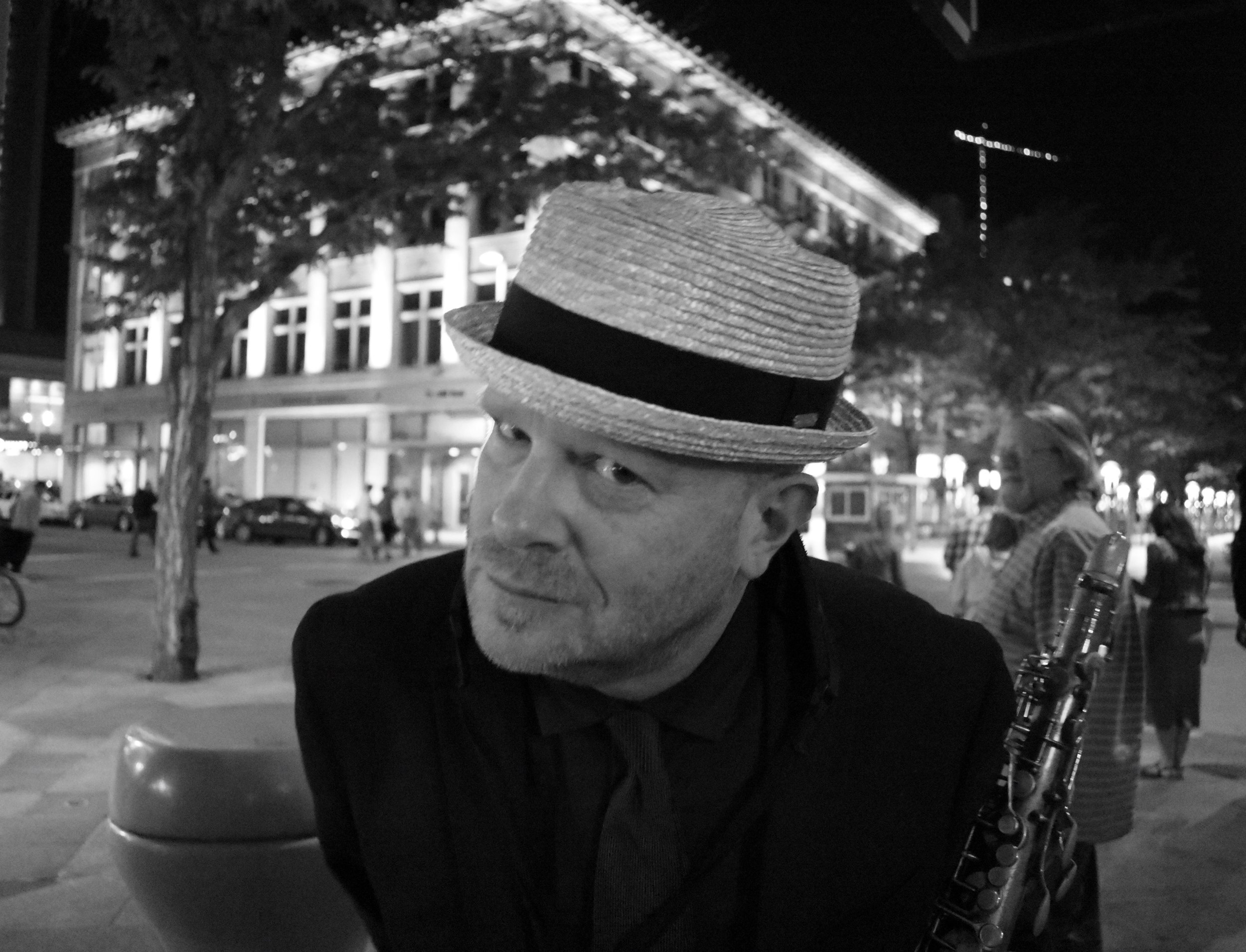 Black and white photo of Jon Hegel, a white man with a black coat, dress shirt and tie, holding a clarinet with a busy night street behind him.