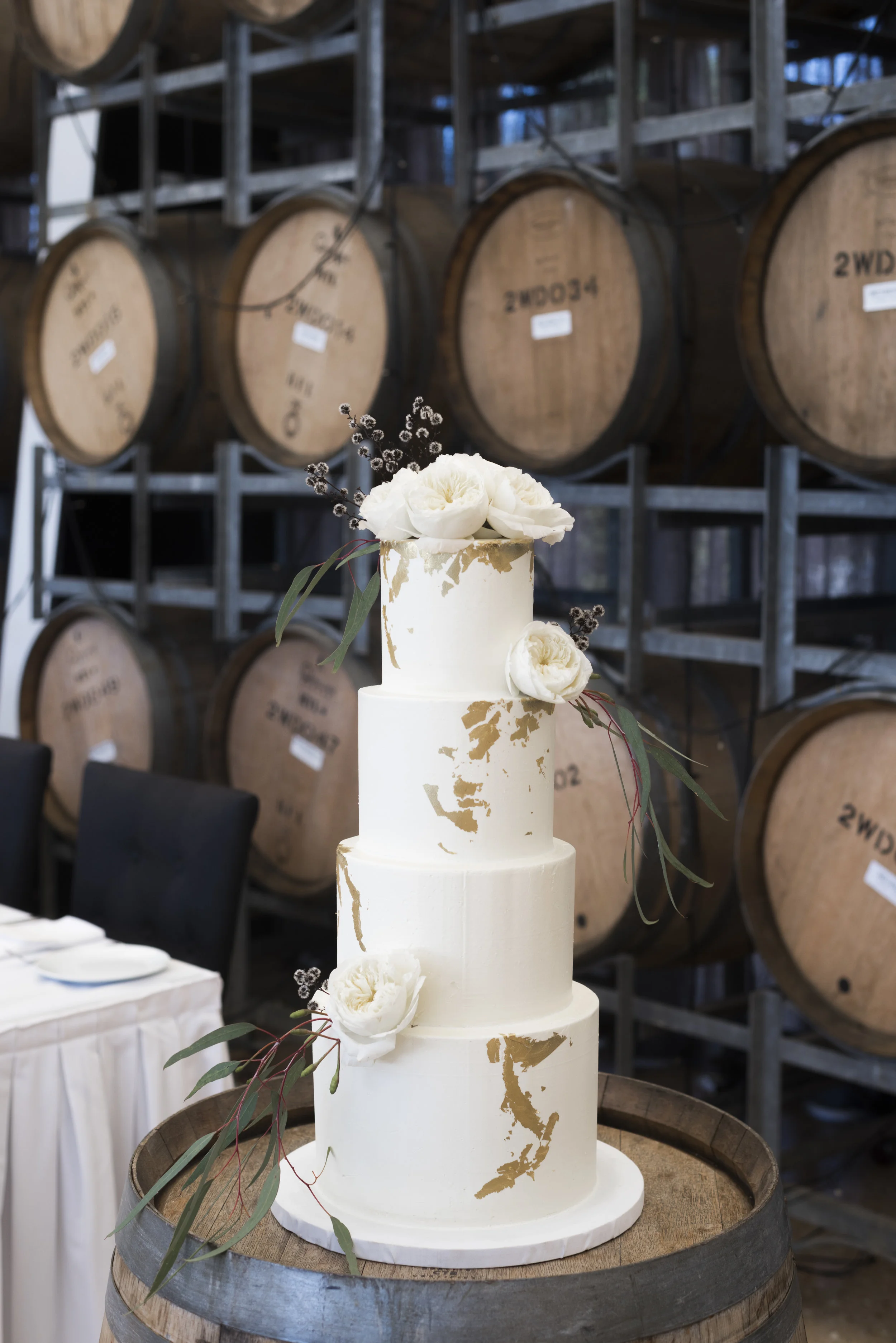 Four tier wedding cake with gold leaf by Edible Art Margaret River . Finished off with cream David Austin roses, gum leaves and strilingia