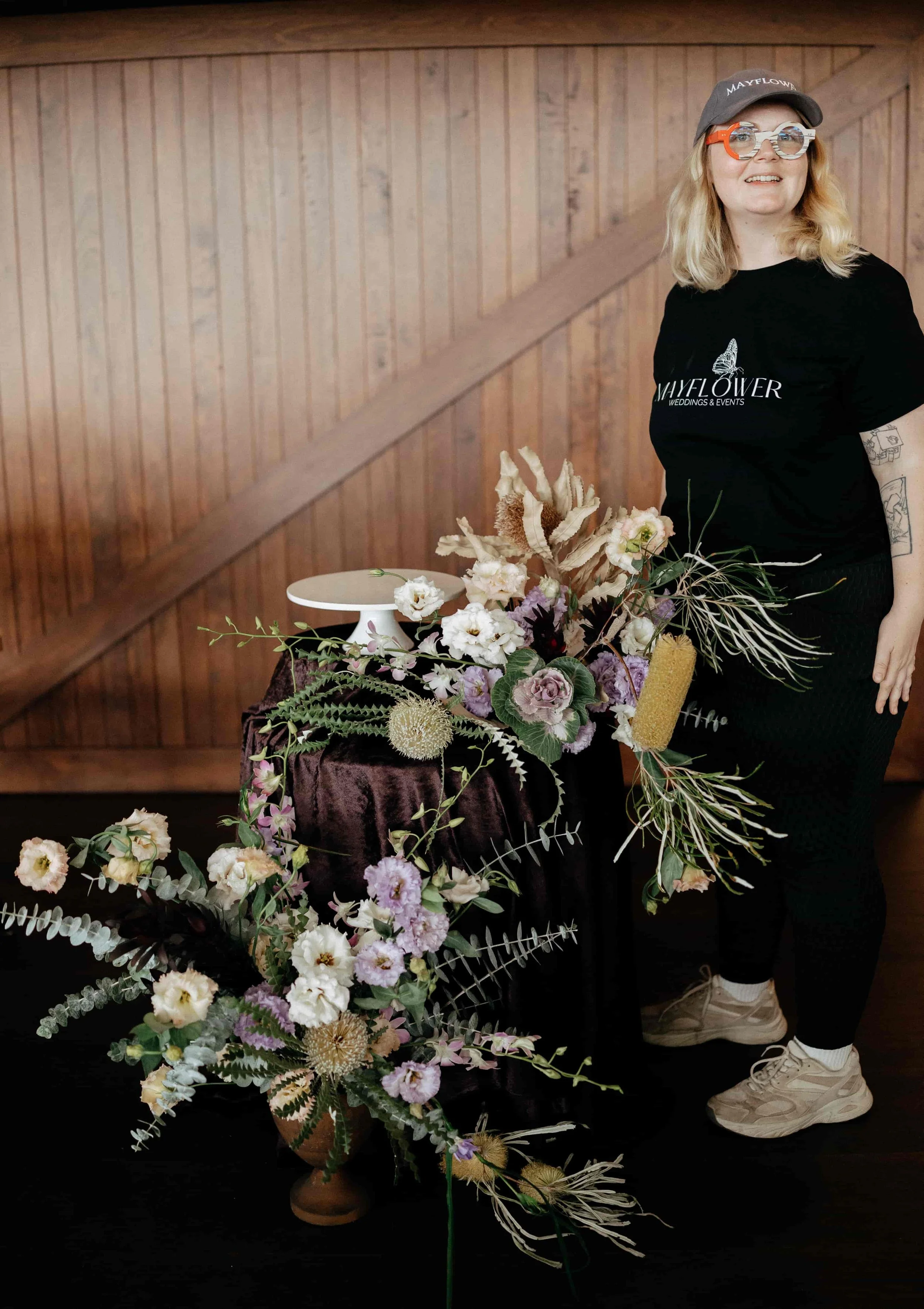 A woman with blonde hair, wearing a black shirt with a butterfly logo, a cap, and sunglasses, standing beside an arrangement of diverse flowers and greenery on a table covered with velvet fabric in front of a wooden wall.
