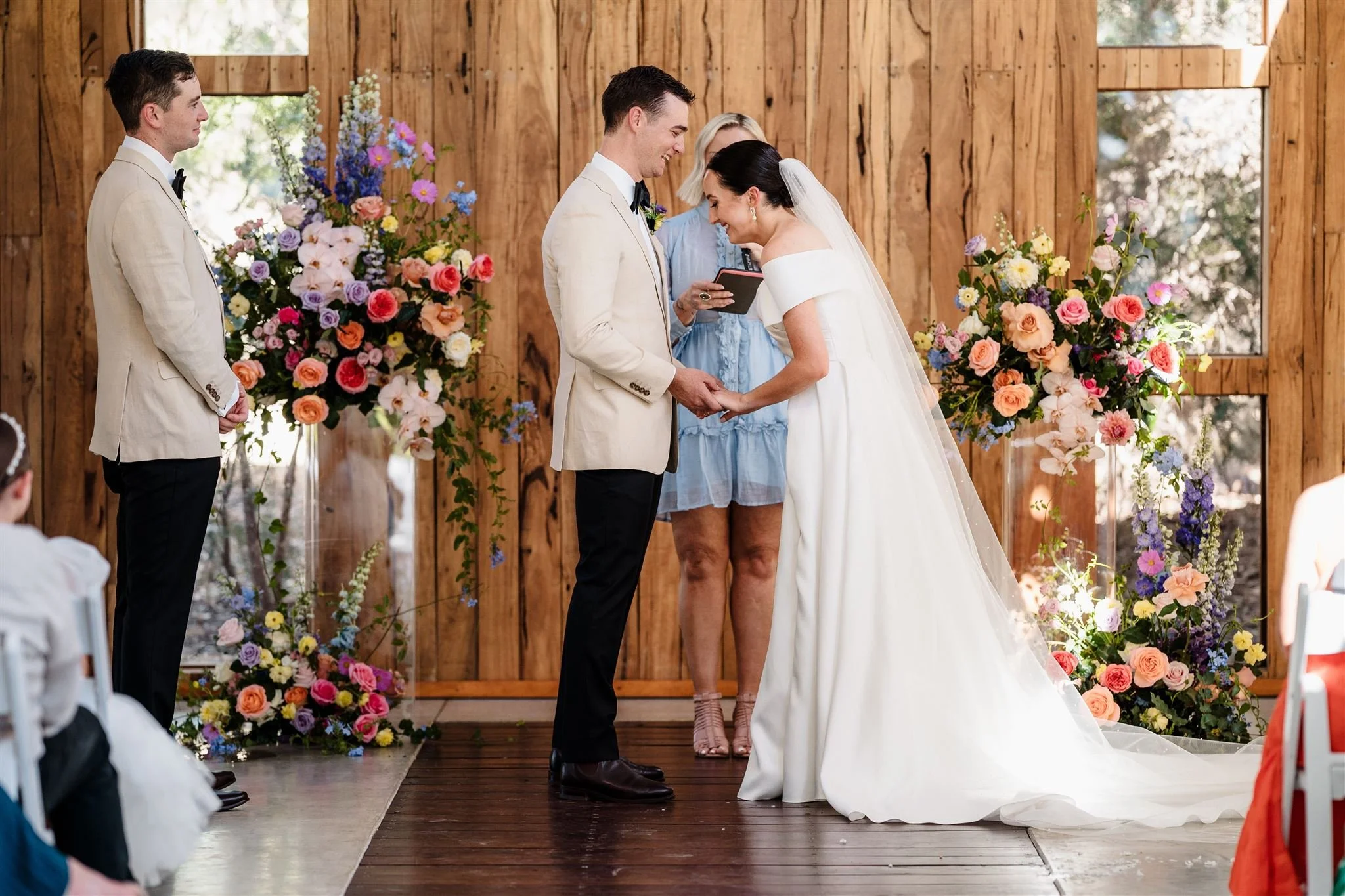 A couple getting married, holding hands and smiling during their wedding ceremony indoors with wooden walls and colorful floral arrangements.