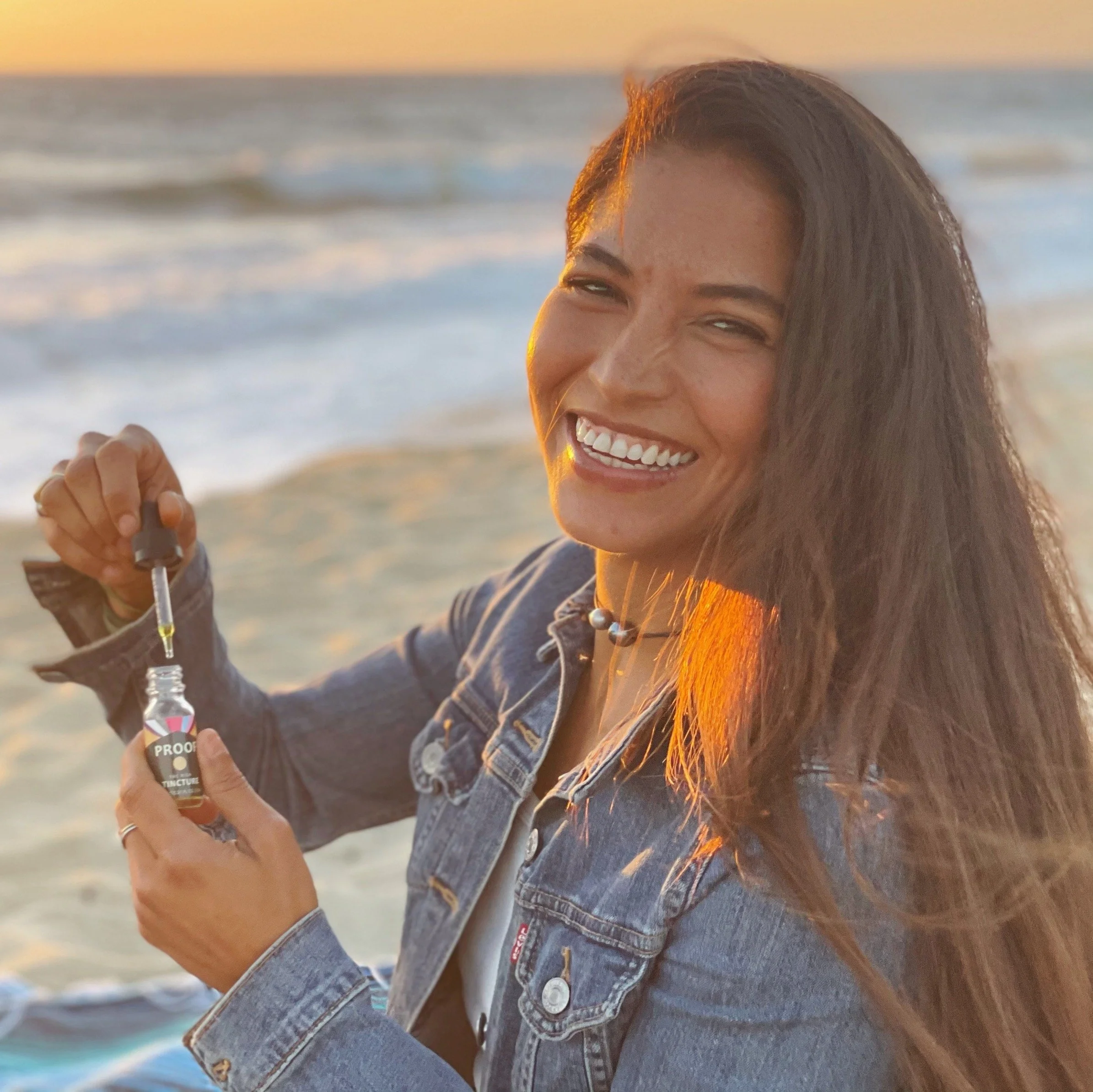 Smiling woman on the beach at sunset holding Proof tincture bottle