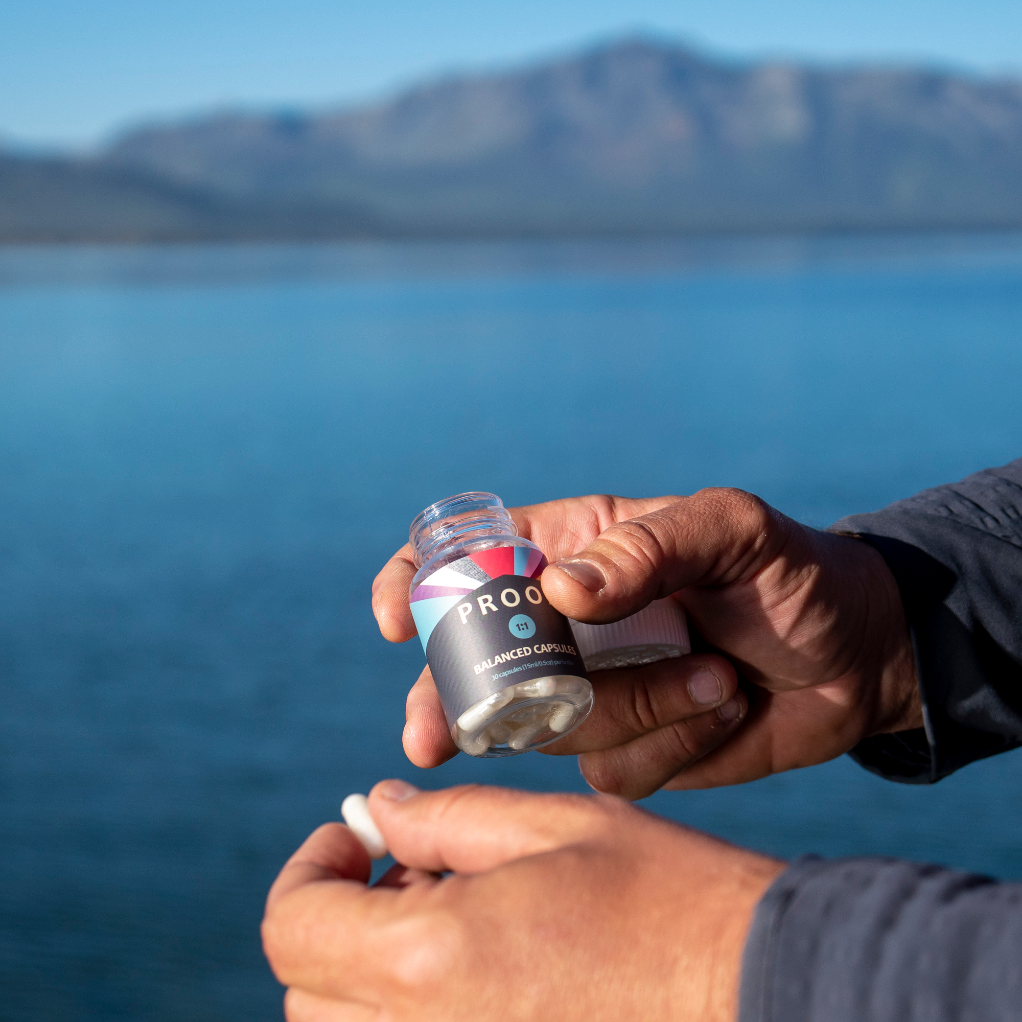 Hands holding PROOF balanced capsules with a scenic lake and mountains in the background.