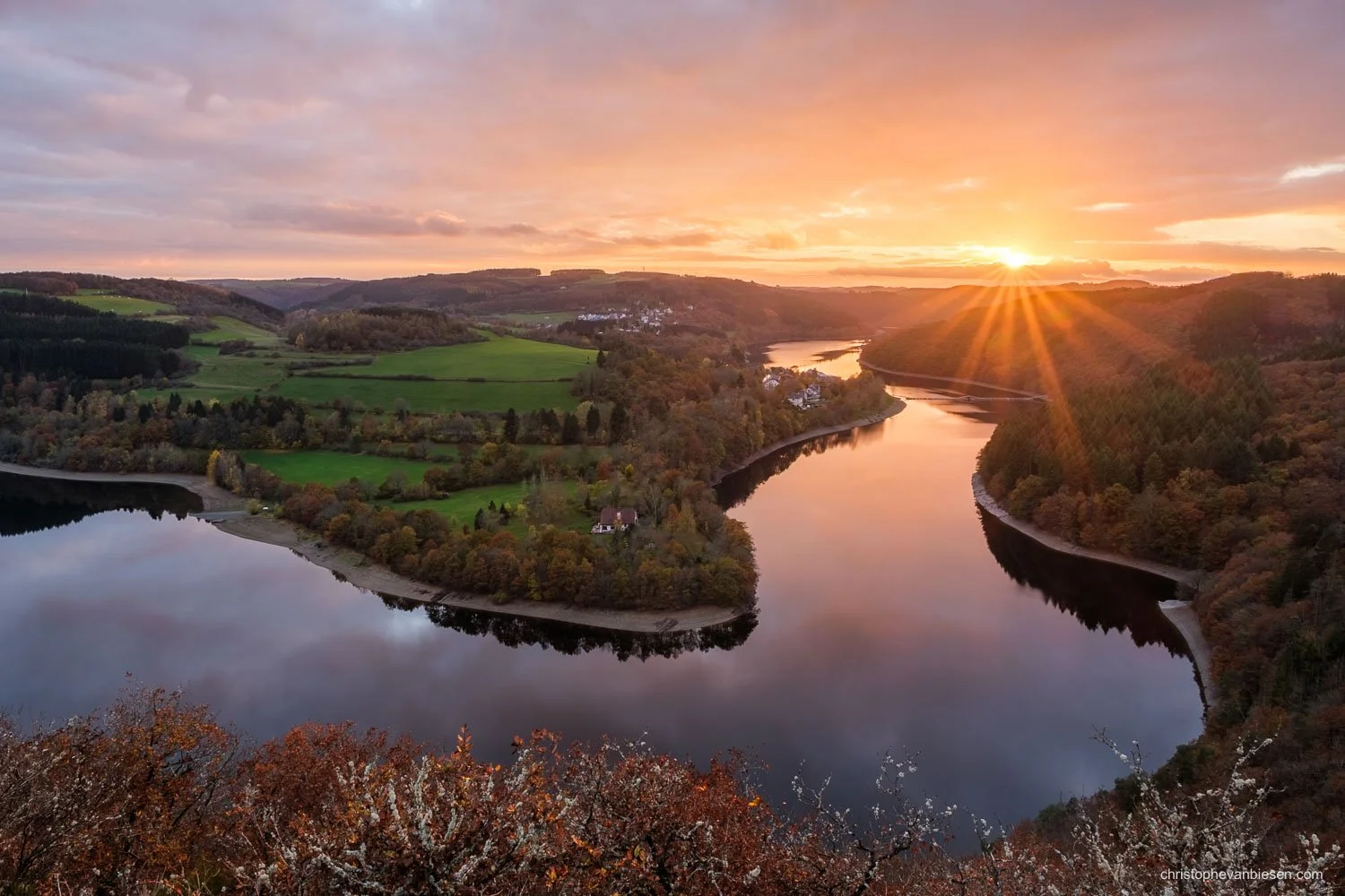 Sunset over a winding lake surrounded by wooded hills, with orange light reflected on the water.