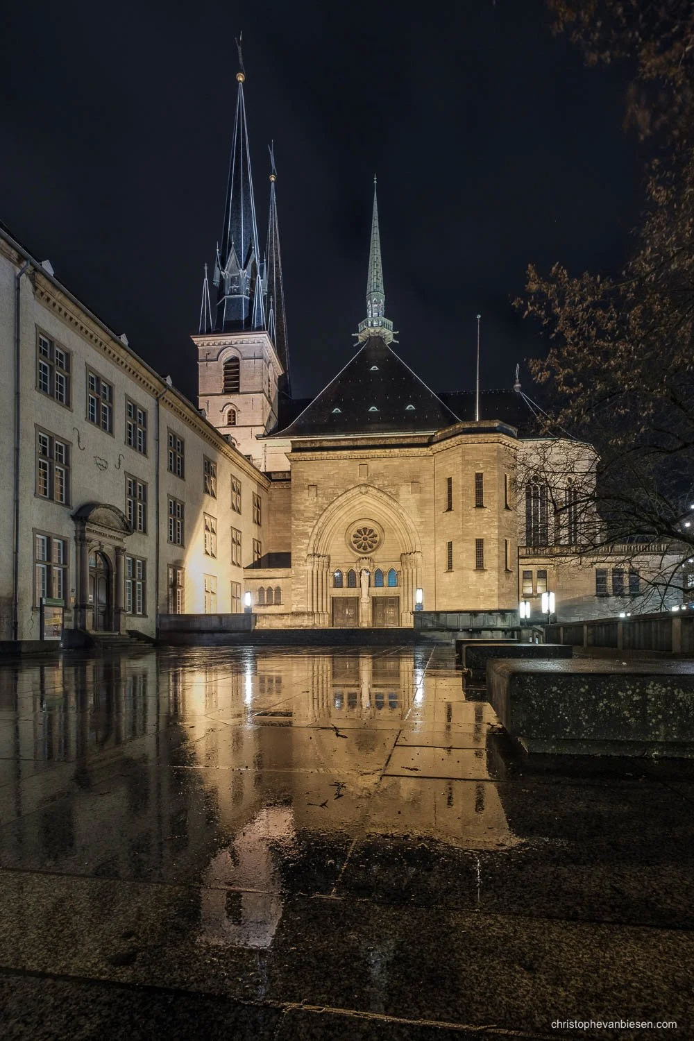Notre-Dame Cathedral in Luxembourg City at night, reflected in wet paving beneath a dark sky.