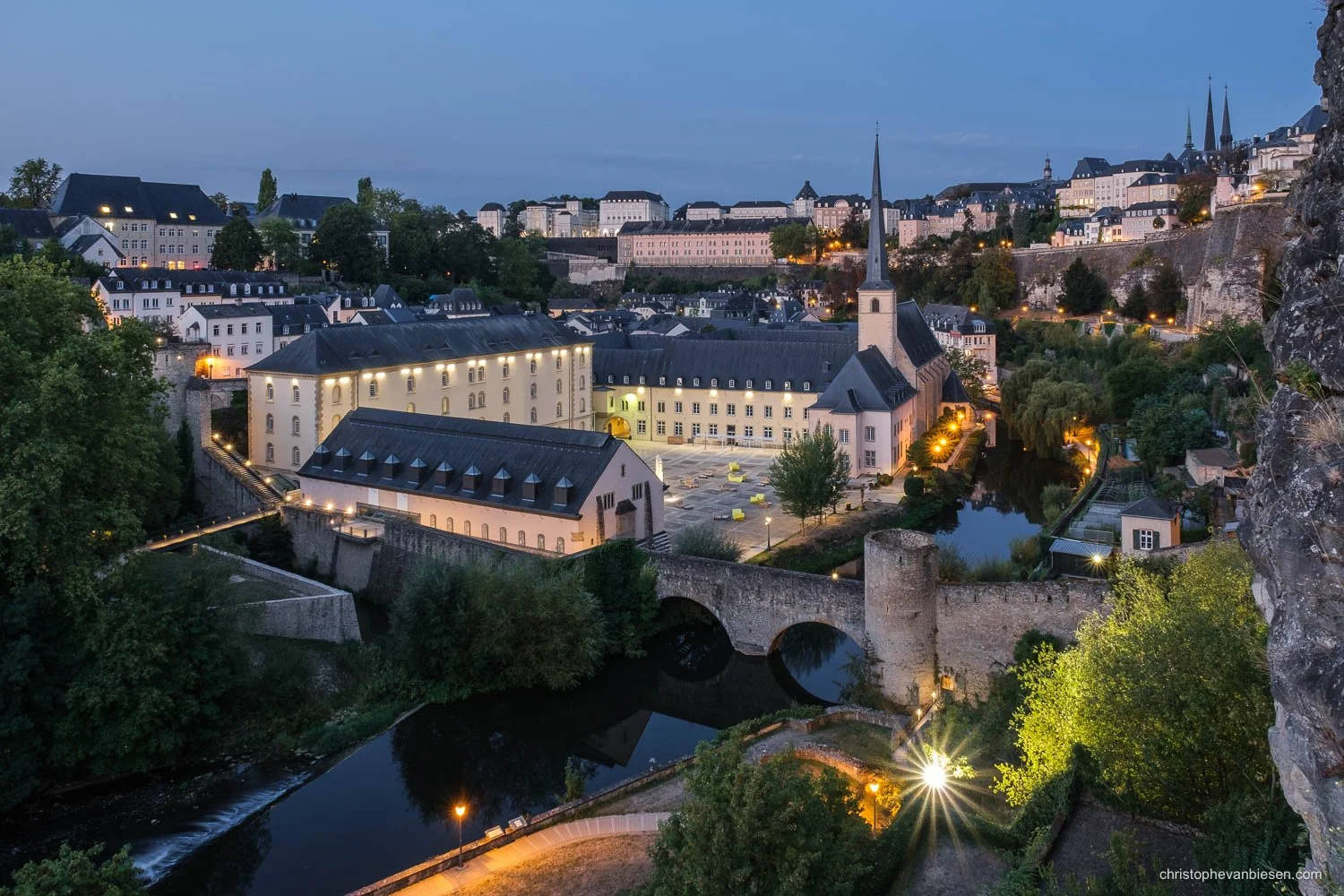 Neumünster Abbey in Luxembourg City at blue hour, with a stone bridge reflected in the Alzette.