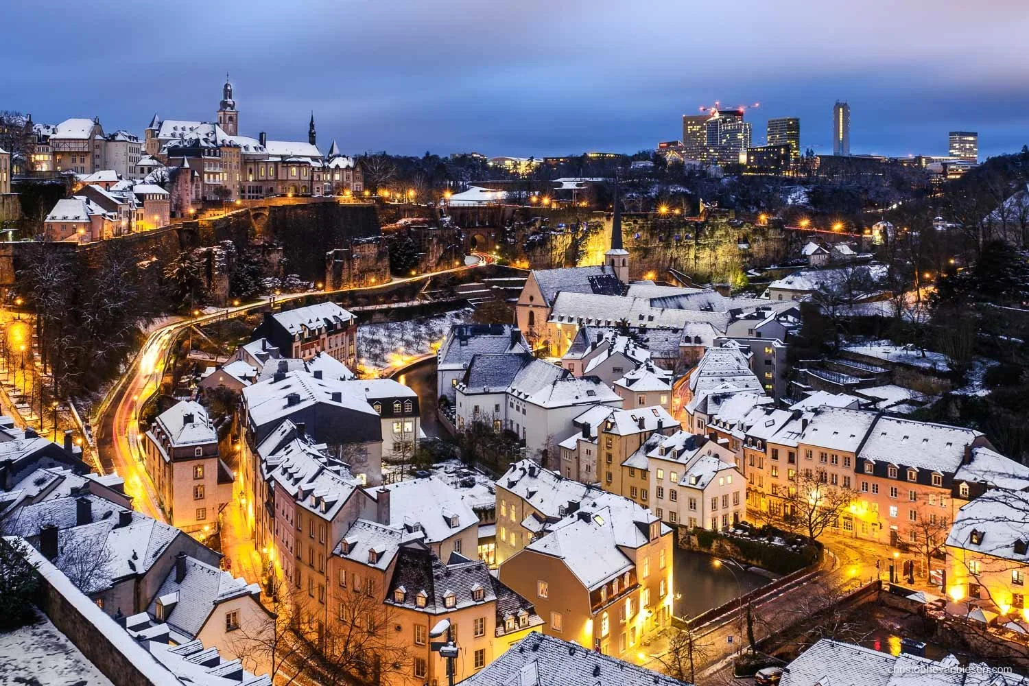 Snow-covered rooftops in Luxembourg’s Grund at dusk, with warm street lights and Kirchberg in the distance.