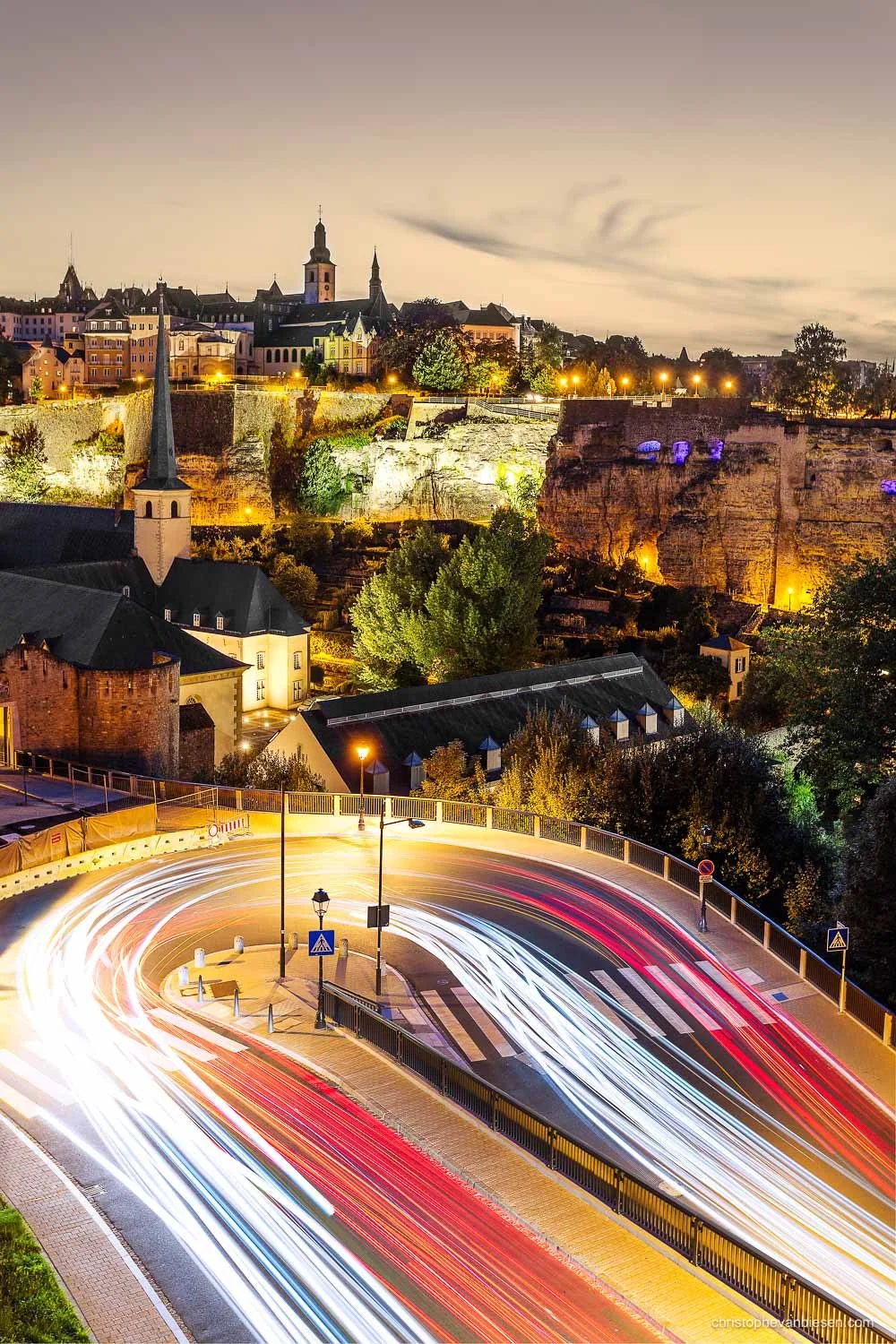 Red and white light trails on a bend above Luxembourg’s Grund, beneath the illuminated old town.