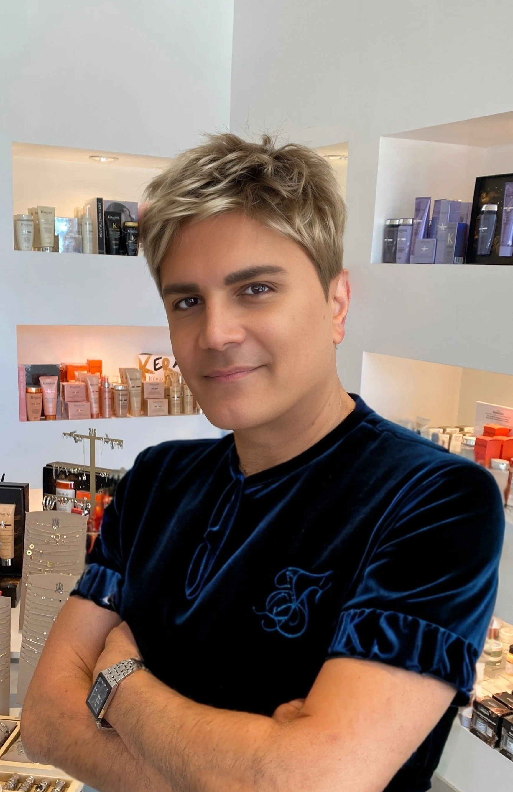 A young man with blonde hair and light skin standing in a beauty product store with arms crossed, wearing a dark velvet shirt.