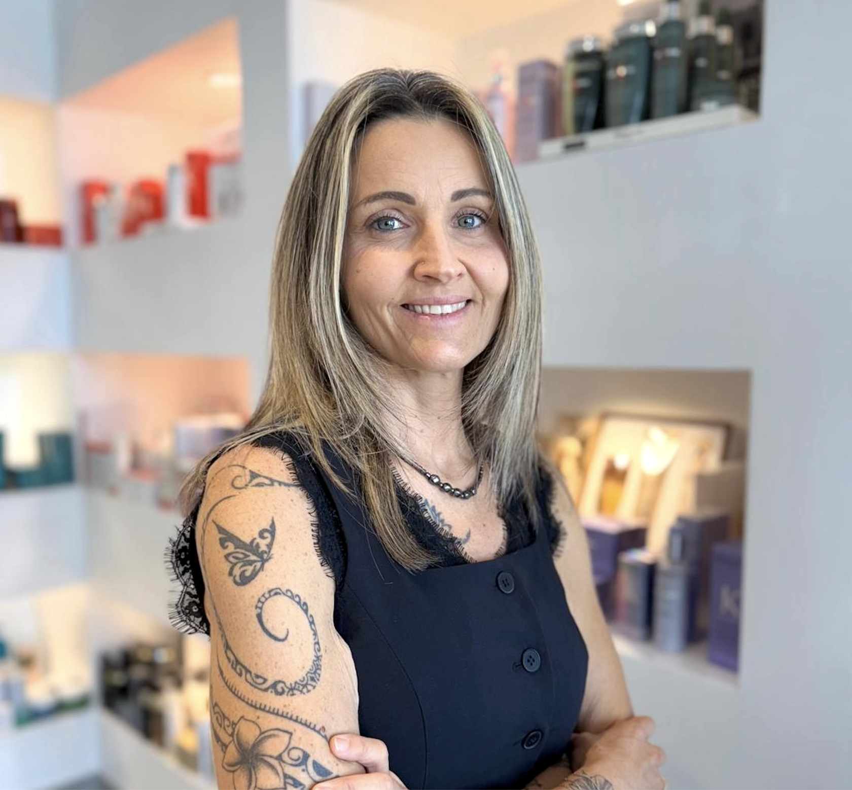 A woman with shoulder-length blonde hair and blue eyes smiling in a store, wearing a black sleeveless dress with lace details on the shoulders and a black beaded necklace, showing tattoos on her left arm.