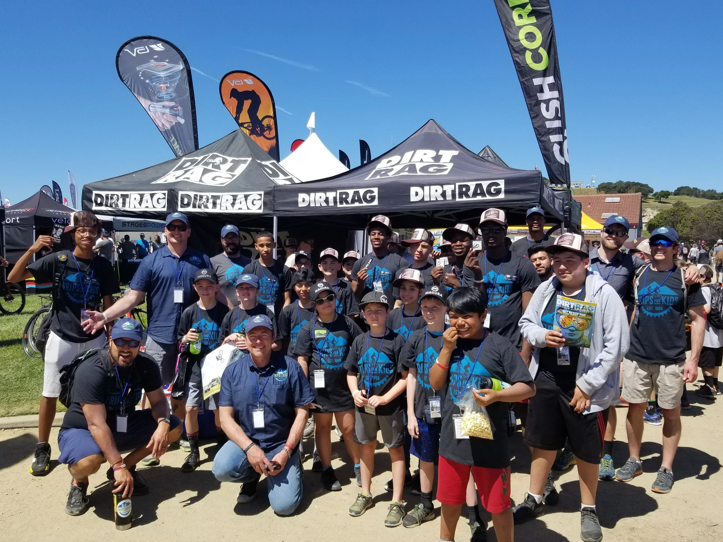 Trips for Kids Executive Director Robert Ping (pictured in first row second from left) joins Trips for Kids participant youth, directors and volunteers in photo in front of sponsor DirtRag Magazine's booth.at the Sea Otter Classic.