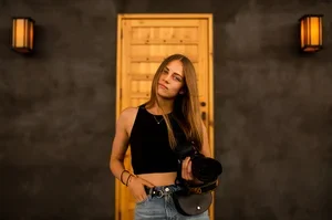 A woman with long brown hair wearing a black crop top and jeans, holding a black hat, standing indoors in front of a wooden door.