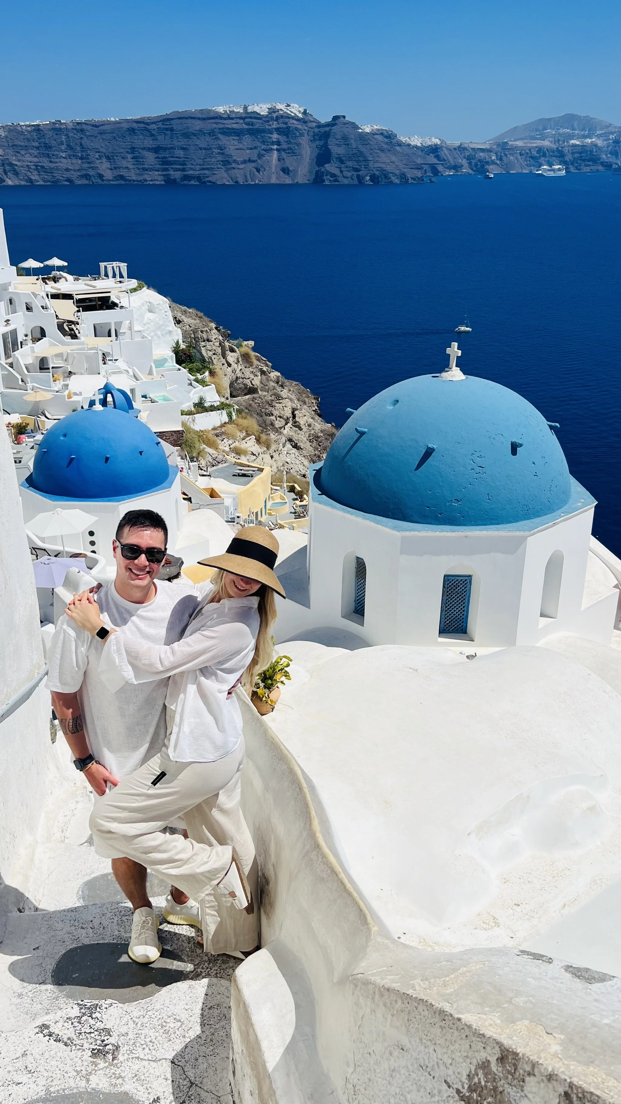 Couple smiling and embracing on a white, curved rooftop in Santorini, Greece, with traditional blue-domed churches and the Aegean Sea in the background.