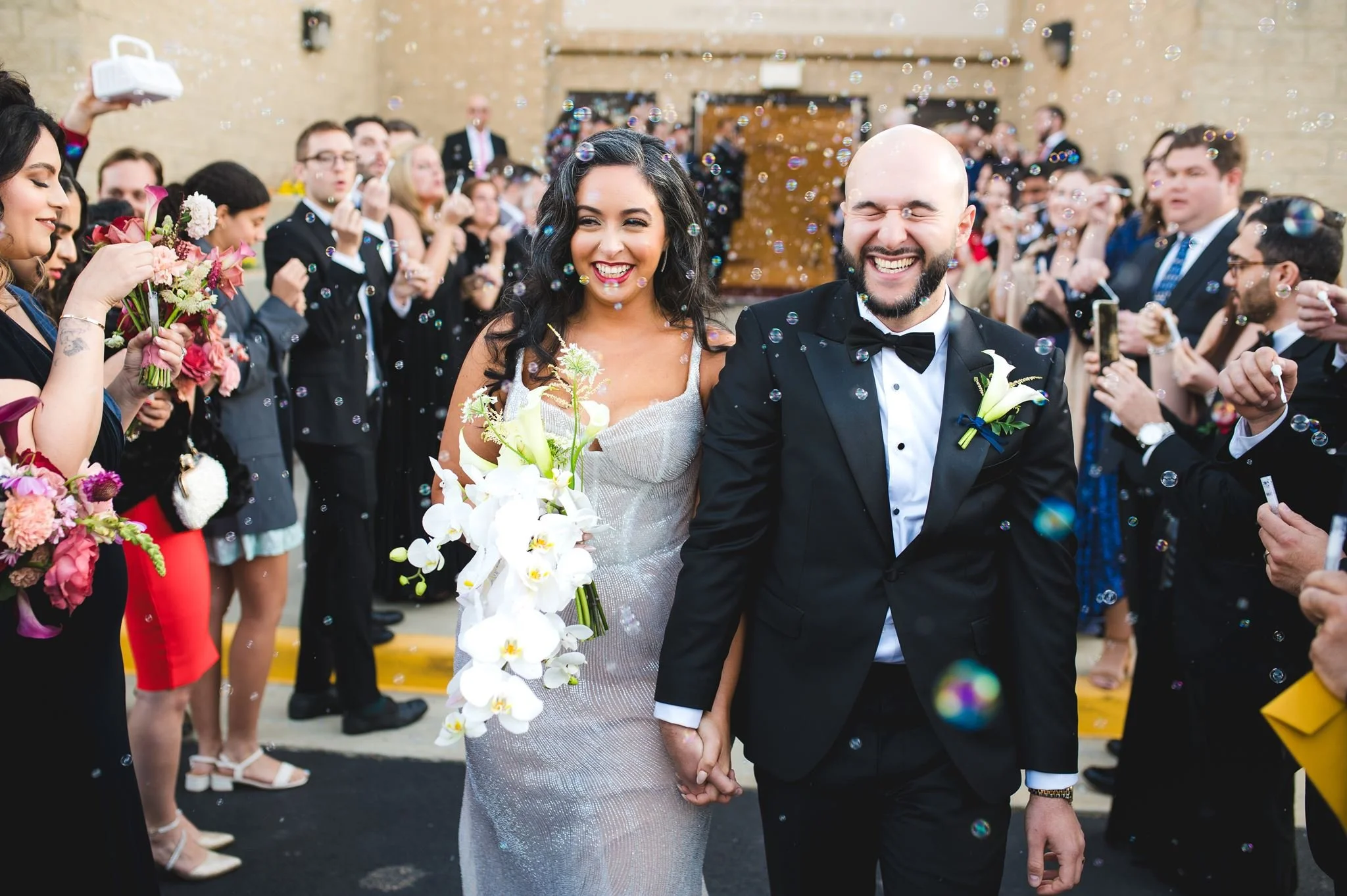 A wedding celebration with a bride and groom holding hands, surrounded by guests and colorful bubbles.