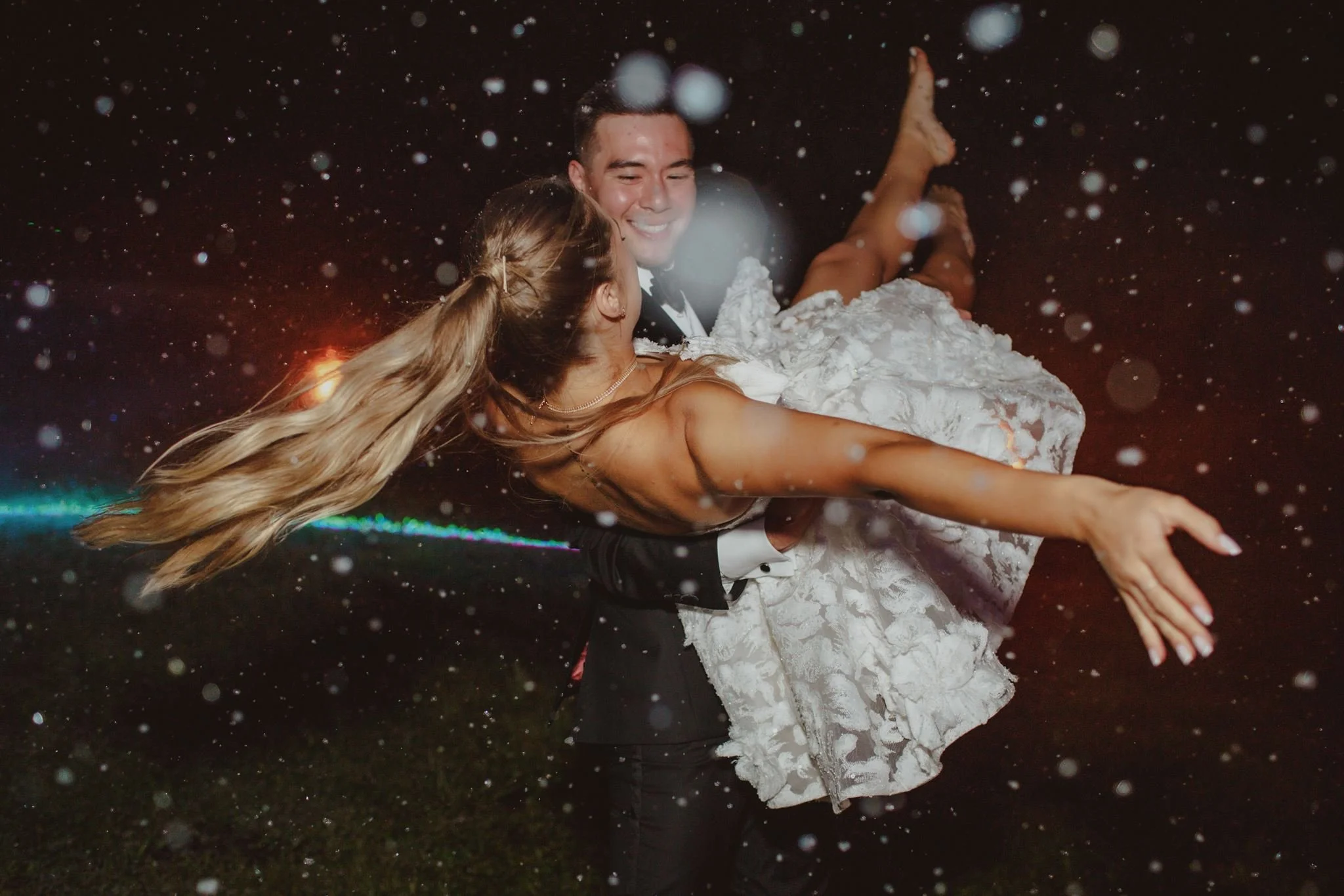 A couple in wedding attire dancing in the rain at night, with the groom smiling and holding the bride, her hair flowing, surrounded by falling rain droplets.