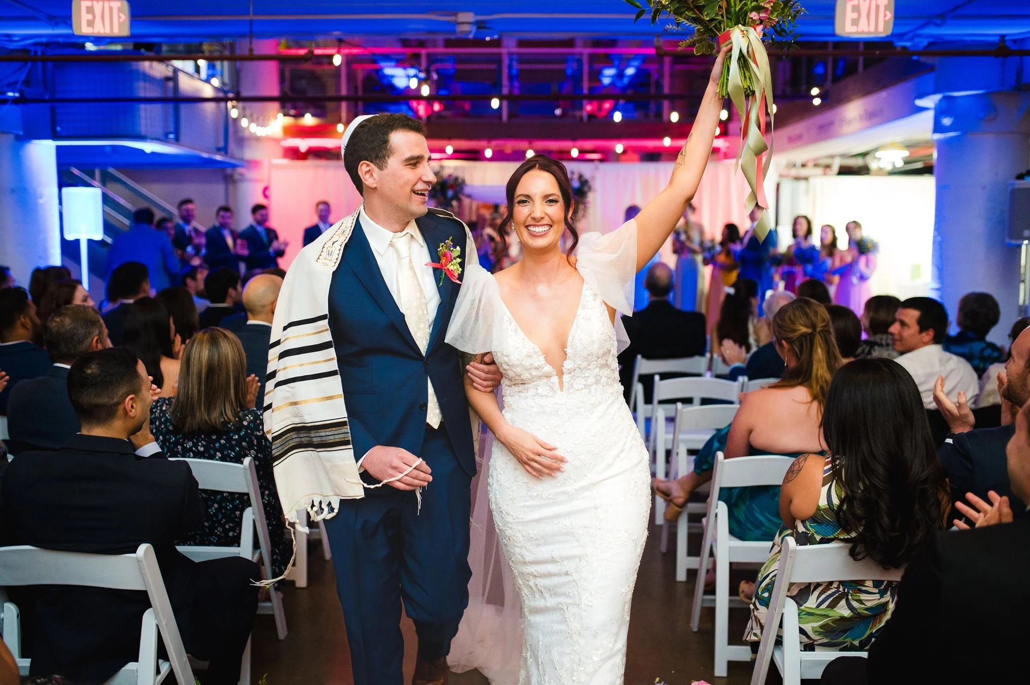 A bride and groom smiling and holding hands during a wedding ceremony, with guests seated in the background and colorful lighting in an indoor venue of the Jewish wedding.