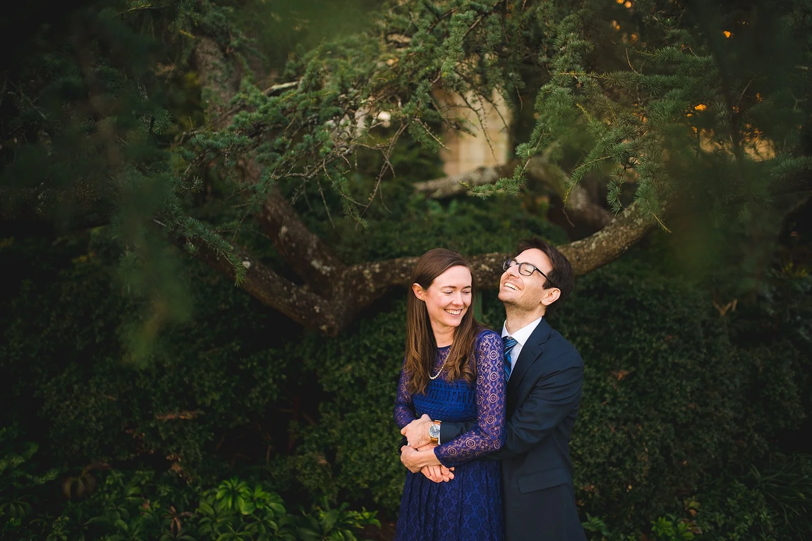 Engaged couple laughing in a hug beneath a cedar tree in Bishop's Garden at Washington National Cathedral, photographed by Gabriele Stonyte Photography