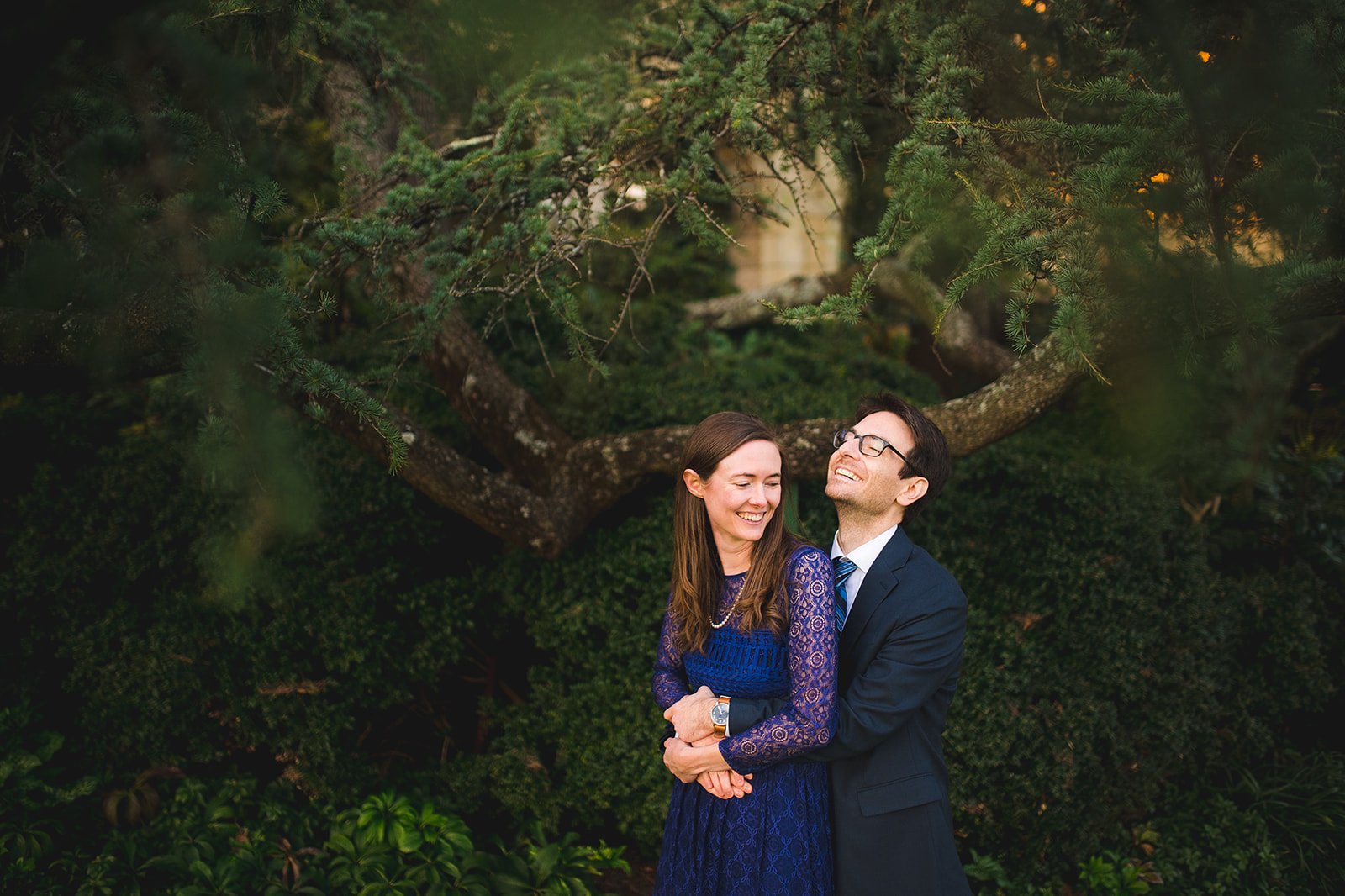 Engaged couple laughing in a hug beneath a cedar tree in Bishop's Garden at Washington National Cathedral, photographed by Gabriele Stonyte Photography