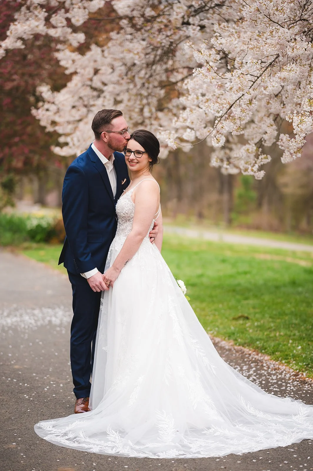 A newlywed couple standing on a pathway under blooming cherry blossom trees, with the groom kissing the bride on her forehead. The bride is wearing a white wedding gown with a long train, and the groom is dressed in a navy suit. Petals are scattered on the ground, and the scene is set outdoors in spring.