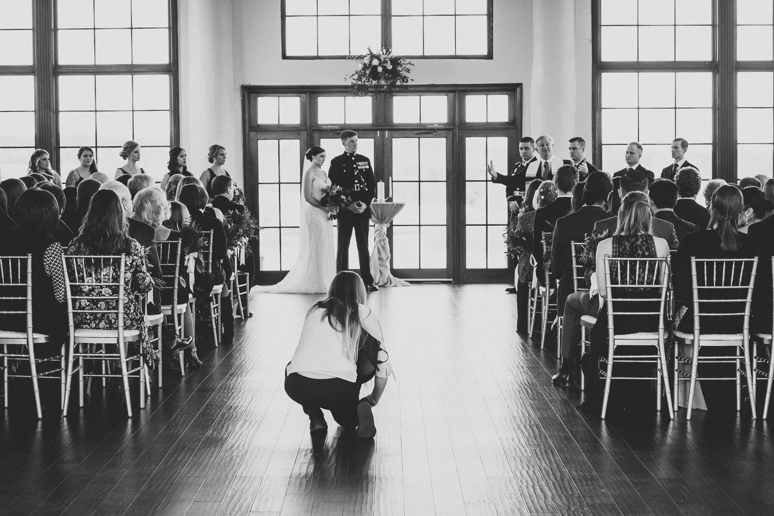 A wedding ceremony taking place indoors with large windows in the background. The bride and groom are standing at the front, with bridesmaids and groomsmen nearby. Guests are seated in chairs on either side. A person in the center is kneeling on the floor, capturing the moment with a camera.