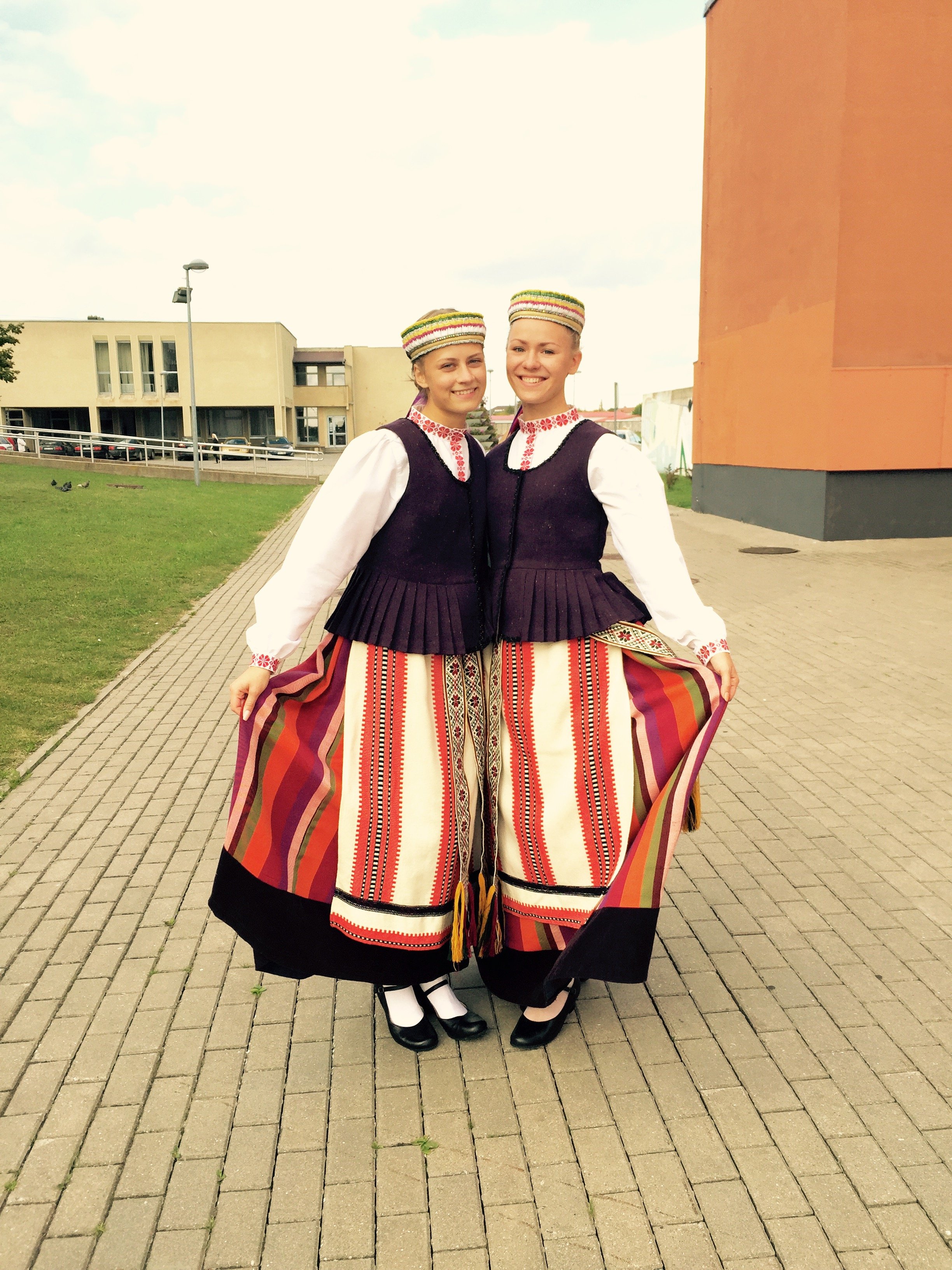 Two women in traditional Baltic folk costumes standing outdoors, smiling, holding their skirts to display colorful striped patterns, with modern buildings and a parking lot in the background.