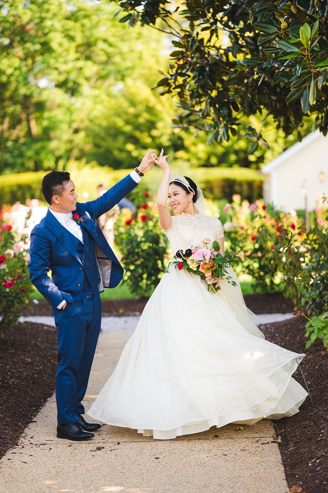 A bride and groom dancing outdoors on a sunny day, surrounded by trees and colorful flowers, with the bride holding a bouquet and wearing a white wedding gown, while the groom wears a blue suit.
