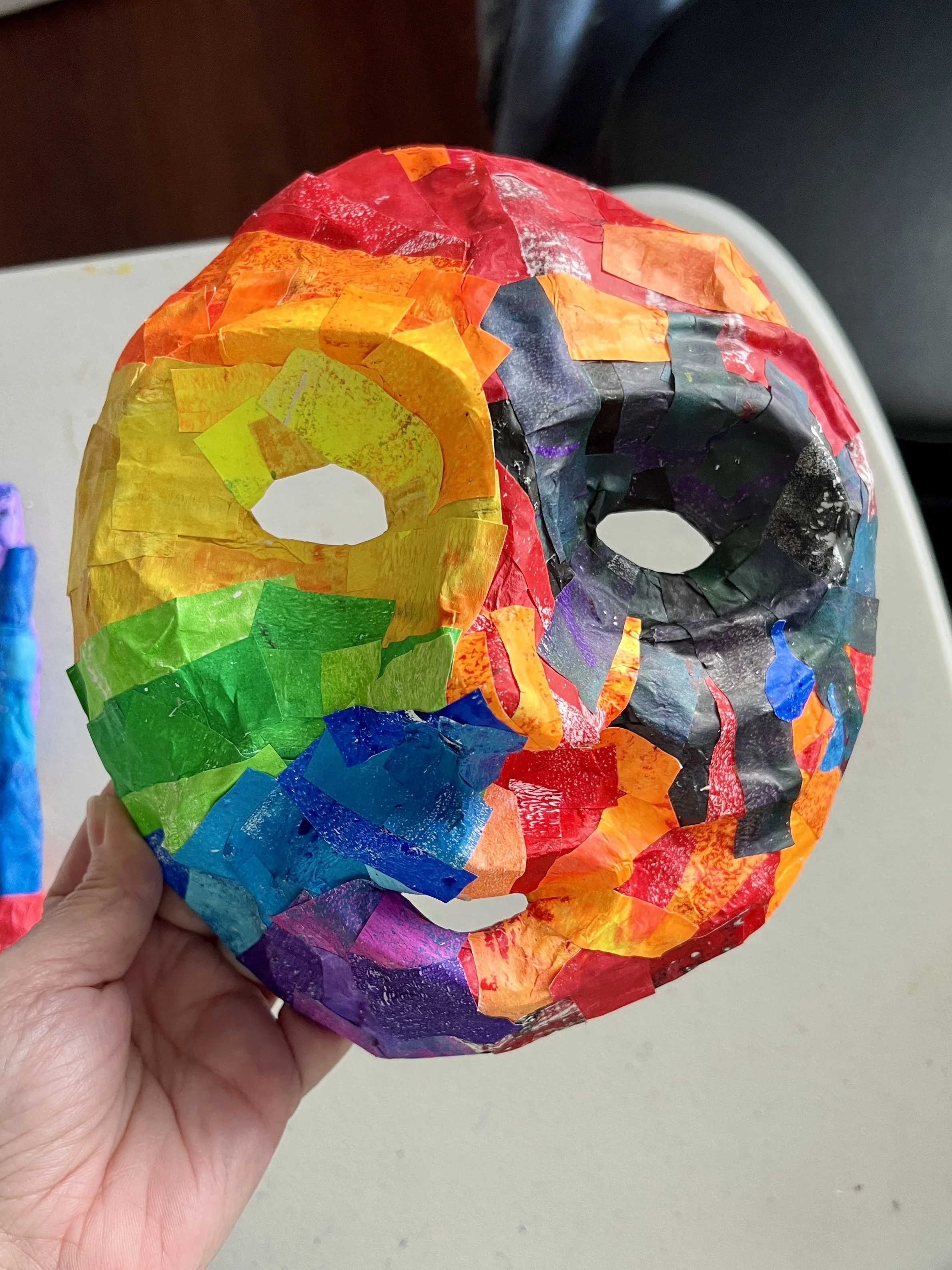 A hand holds a mask that appears to be some time of paper mache or similar. It has a collaged surface made of multiple bright colors. The right eye is black. the mask has a happy expression on one side and an angry on the other.