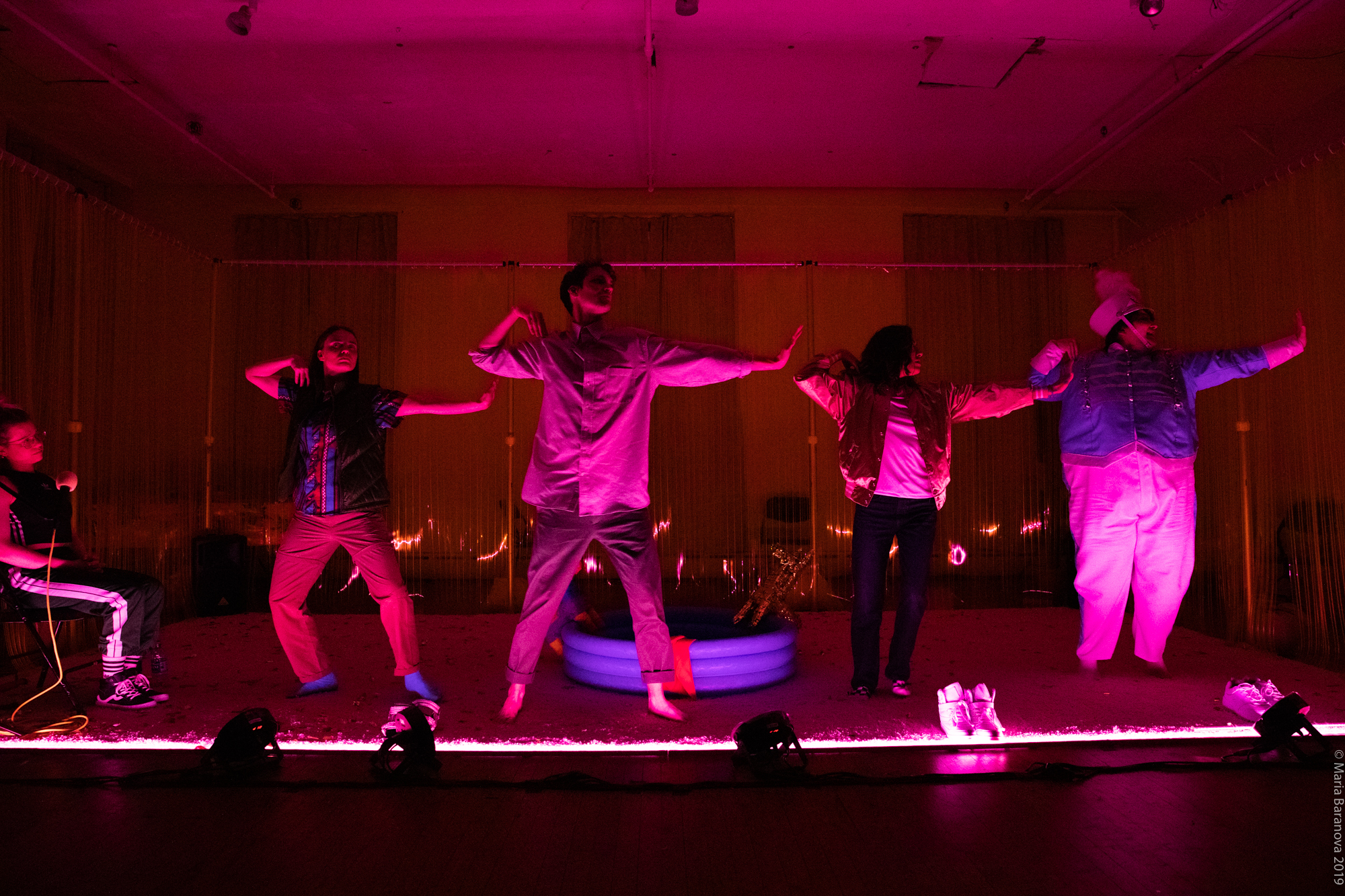 a row of four humans posing as if in a choreographed dance, they are lit from below in a vibrant pink glow which fills the room