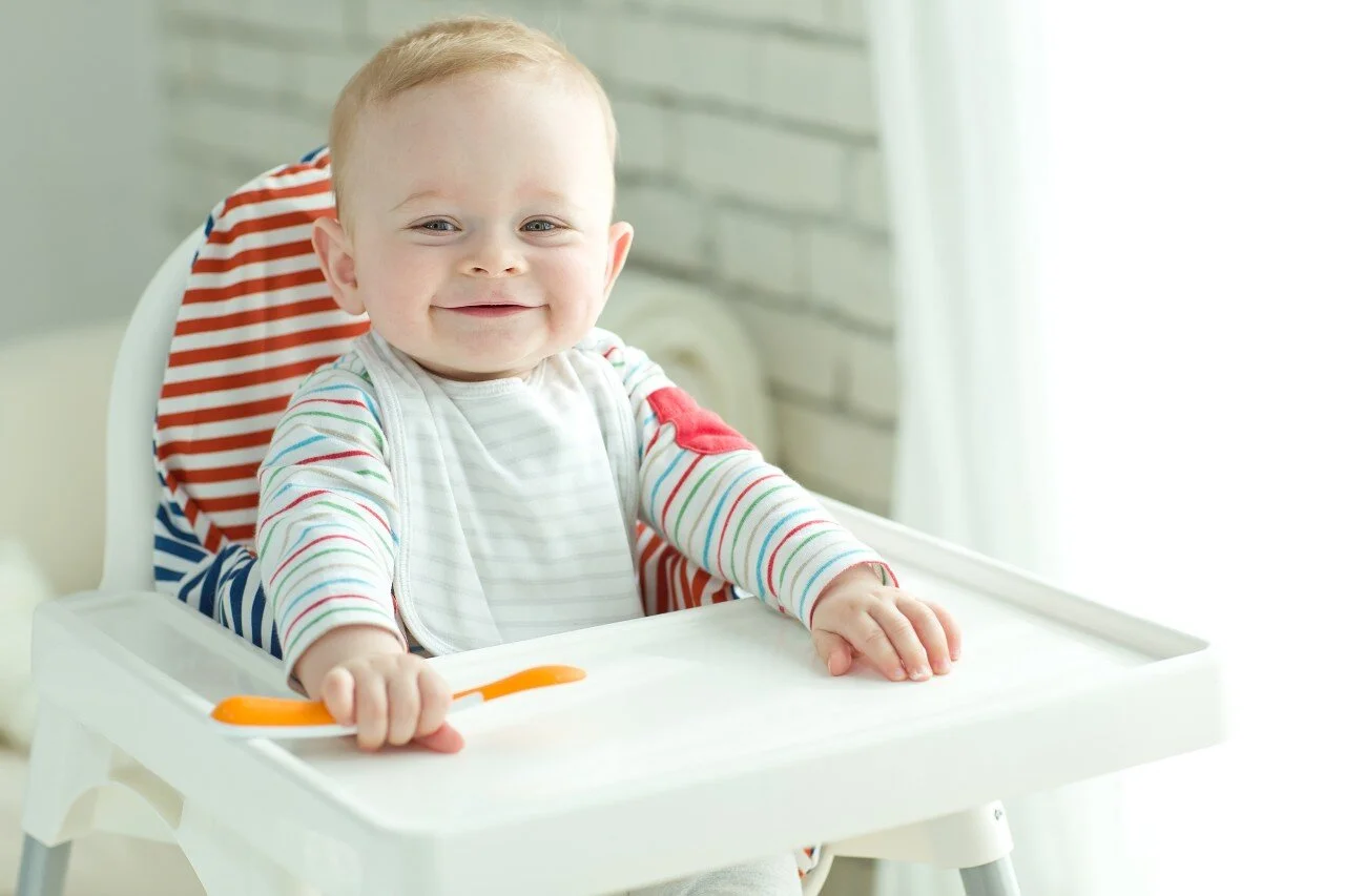 Baby boy sat in white highchair with orange baby spoon