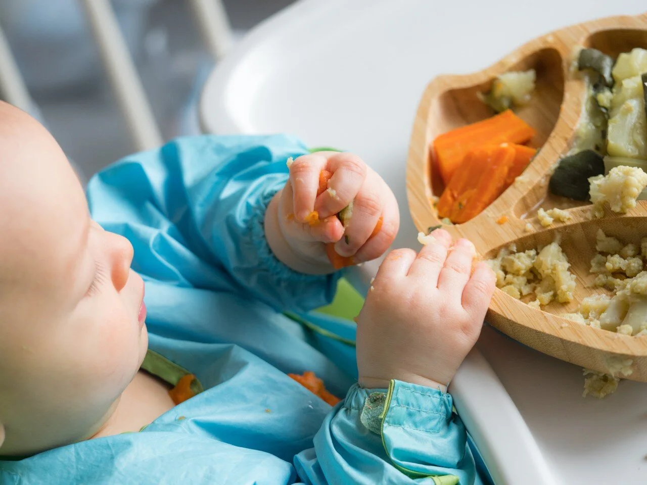 Baby feeding self in highchair wearing a blue bib