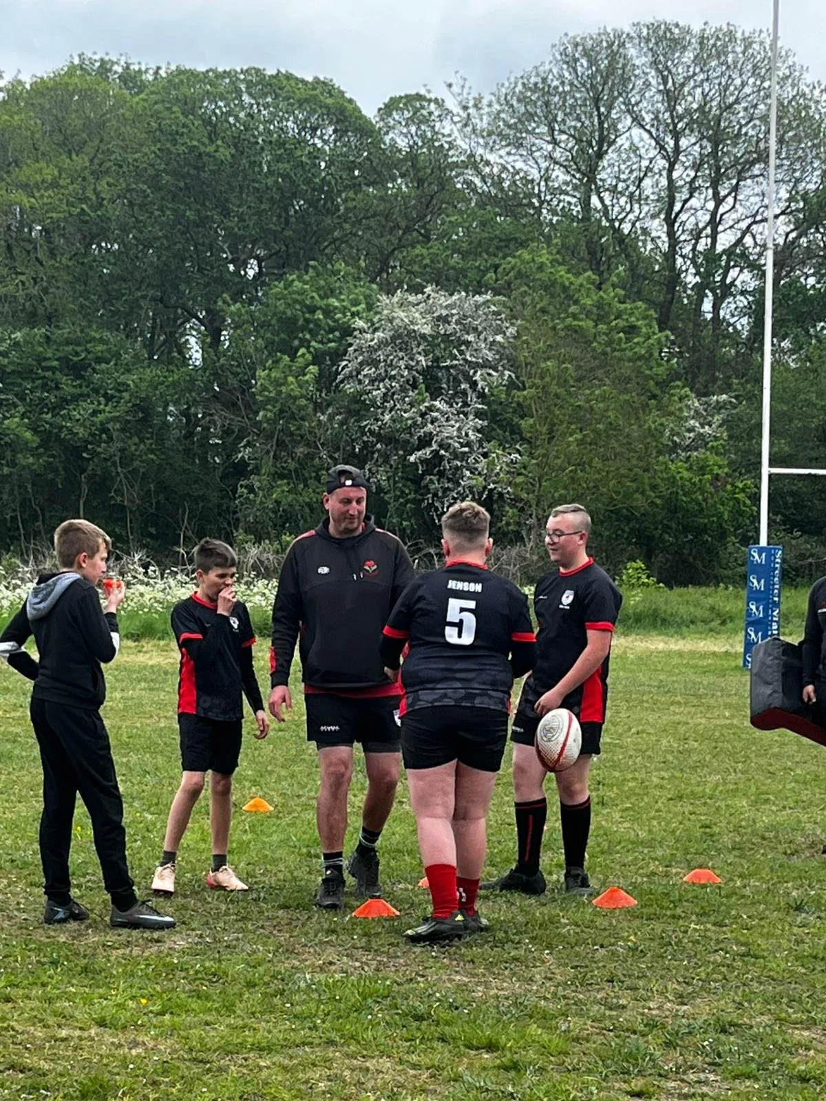 A group of children and a coach on a rugby field during practice. The coach, wearing a black hoodie, is talking to the children who are wearing black and red rugby uniforms. The kids are standing around orange cones, and a rugby ball is visible. Back