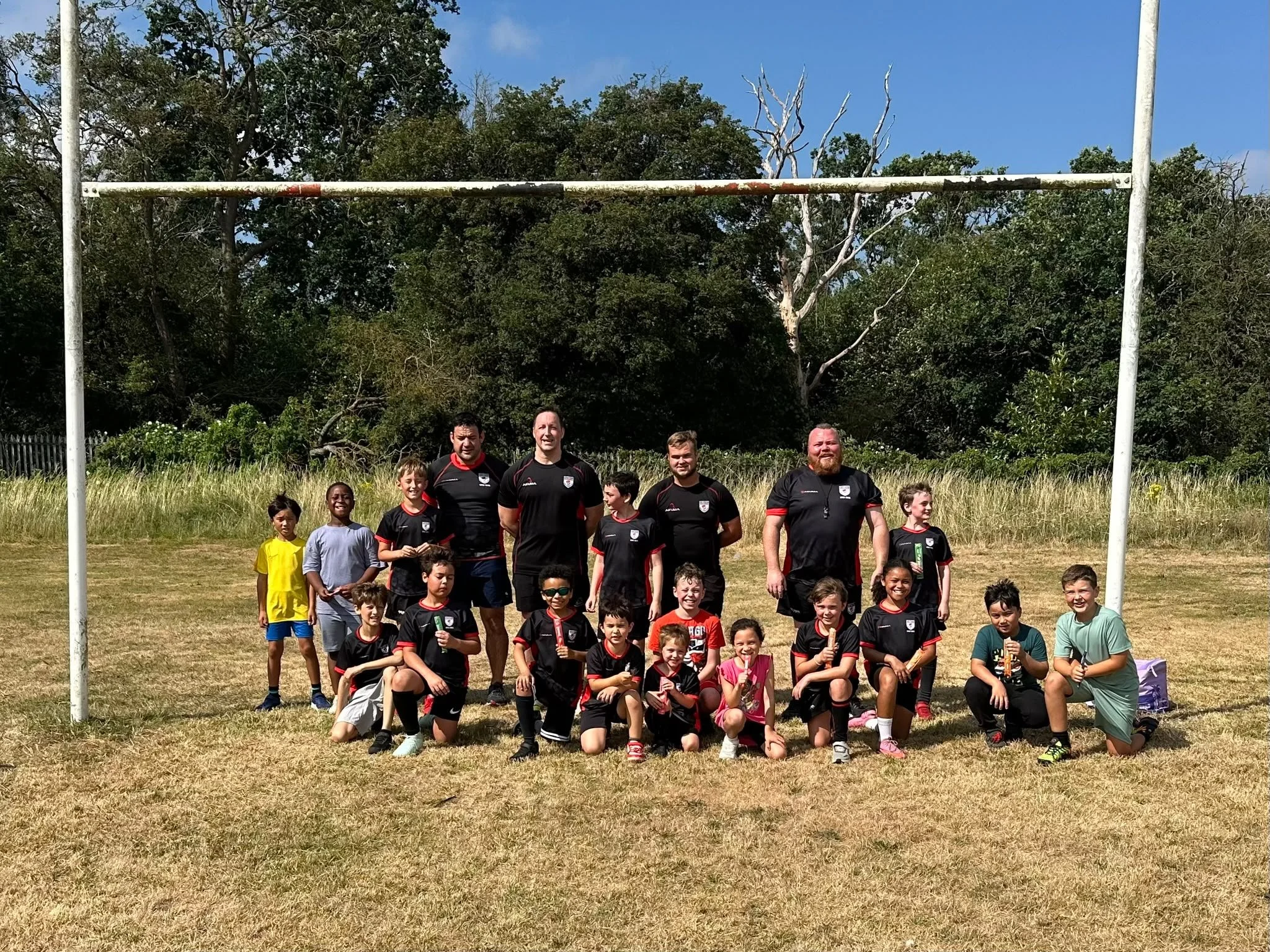 A group of children and adult rugby players are posing for a photo under a rugby goalpost in a grassy field, with trees and a blue sky in the background.