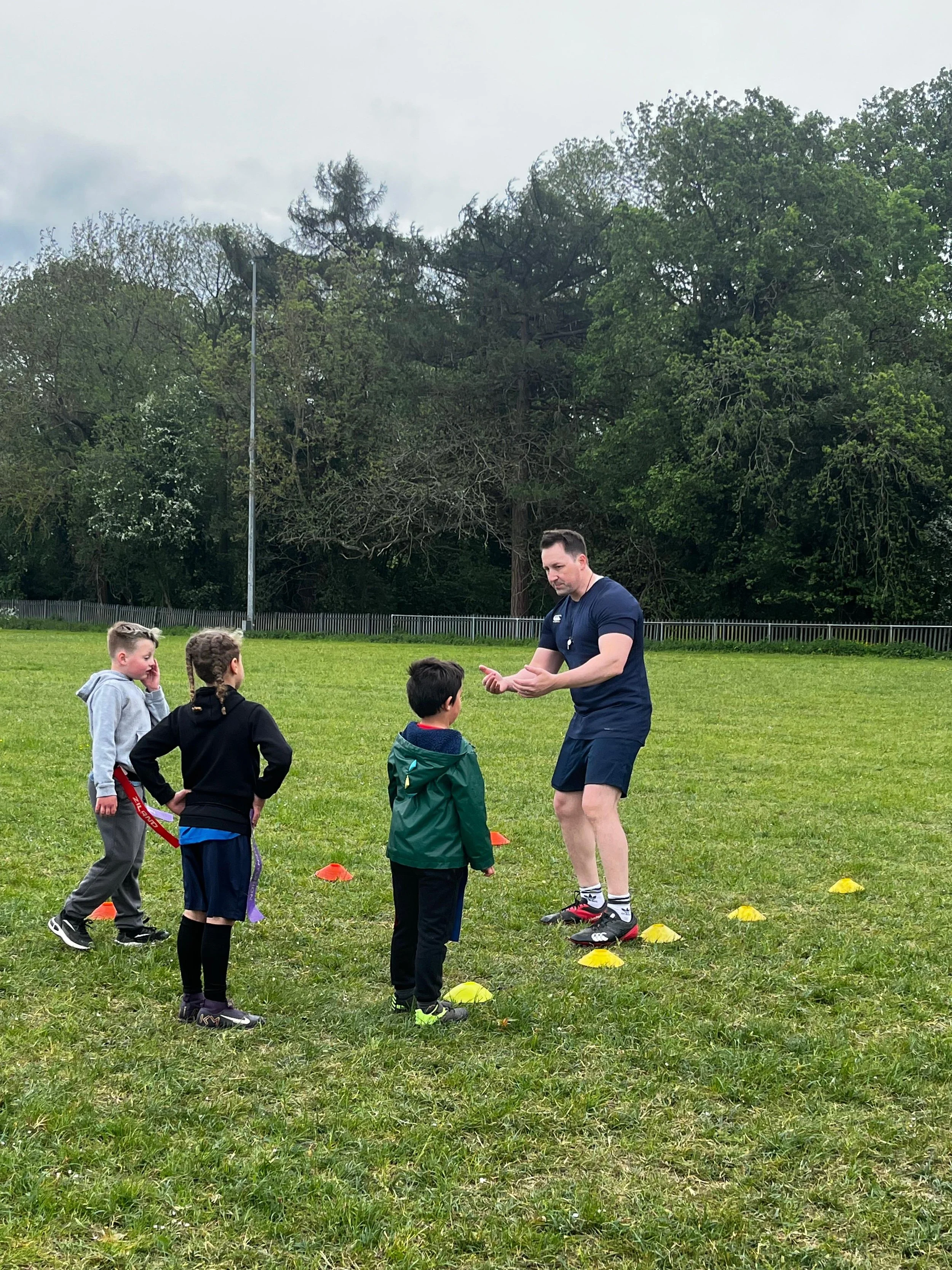 A man leading a fitness training session with children on a grassy field, with trees and a fence in the background.