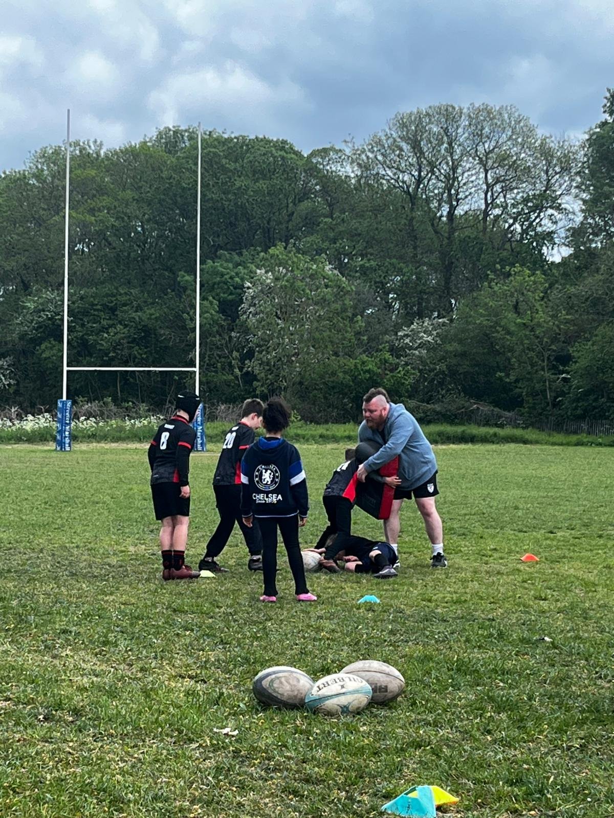 Coaching session with young rugby players on a field, coach assisting a child in tackling drill, rugby balls on grass, trees and cloudy sky in background.
