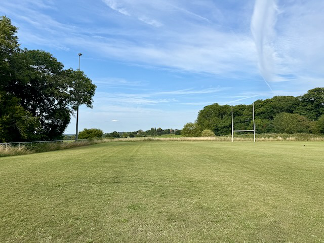 Empty rugby field with goalposts, surrounded by trees under a blue sky with clouds.