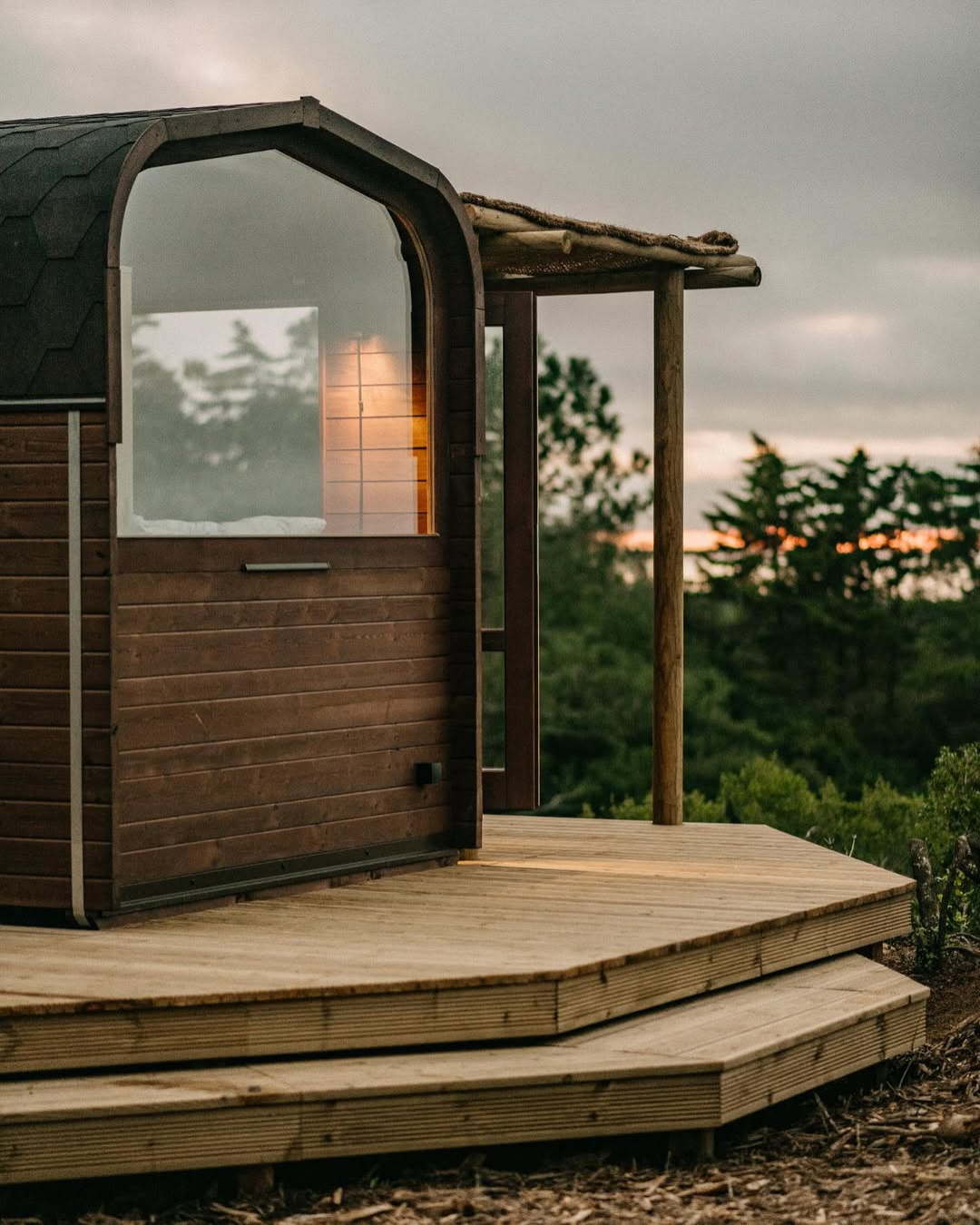 Wooden cabin with arched glass window and timber deck set among pine trees at dusk, Tiny House Escape Portugal