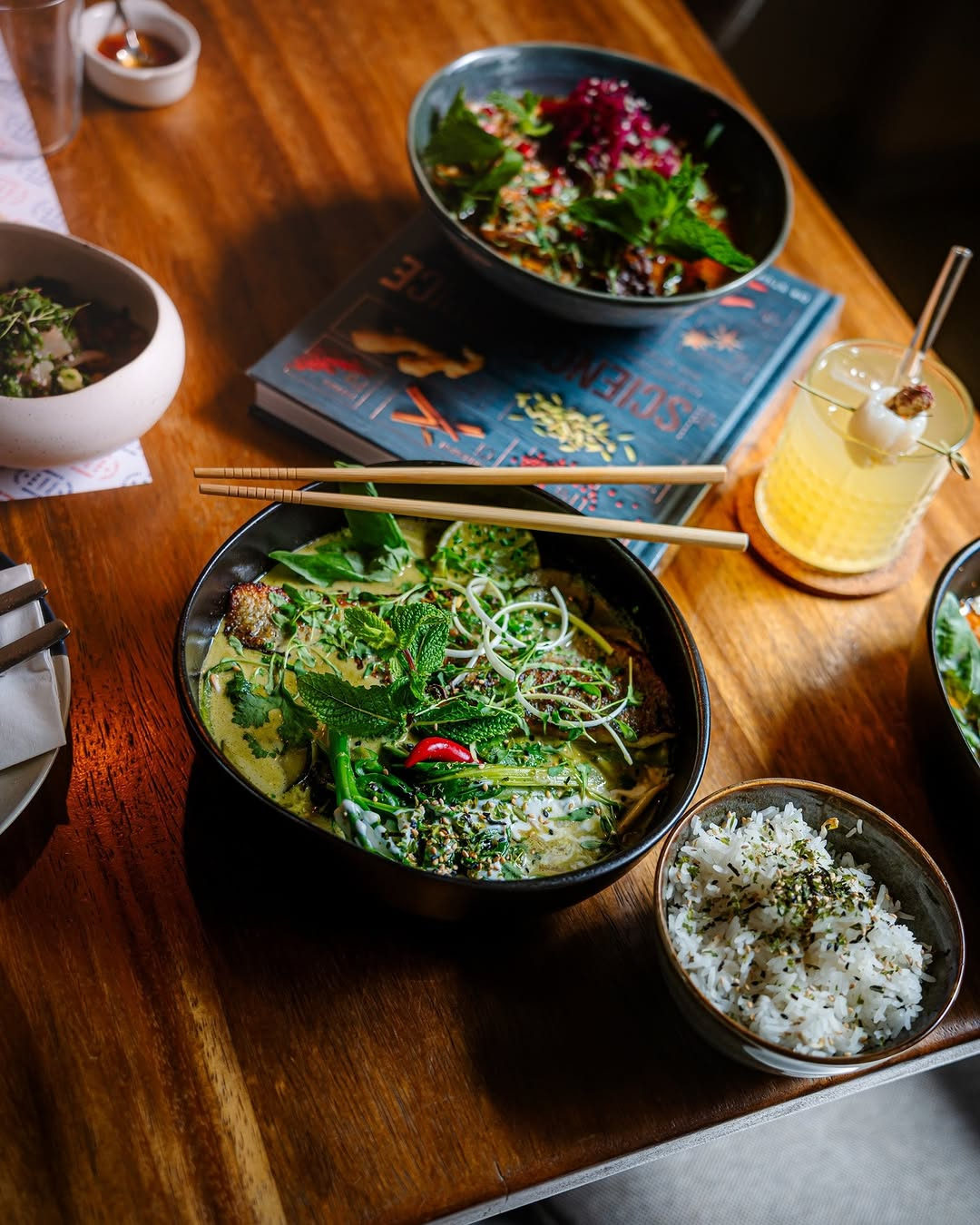 Overhead spread of dishes at Luka Lisboa featuring a green curry bowl with fresh herbs, steamed rice, a pomegranate salad and a cocktail on a warm wooden table