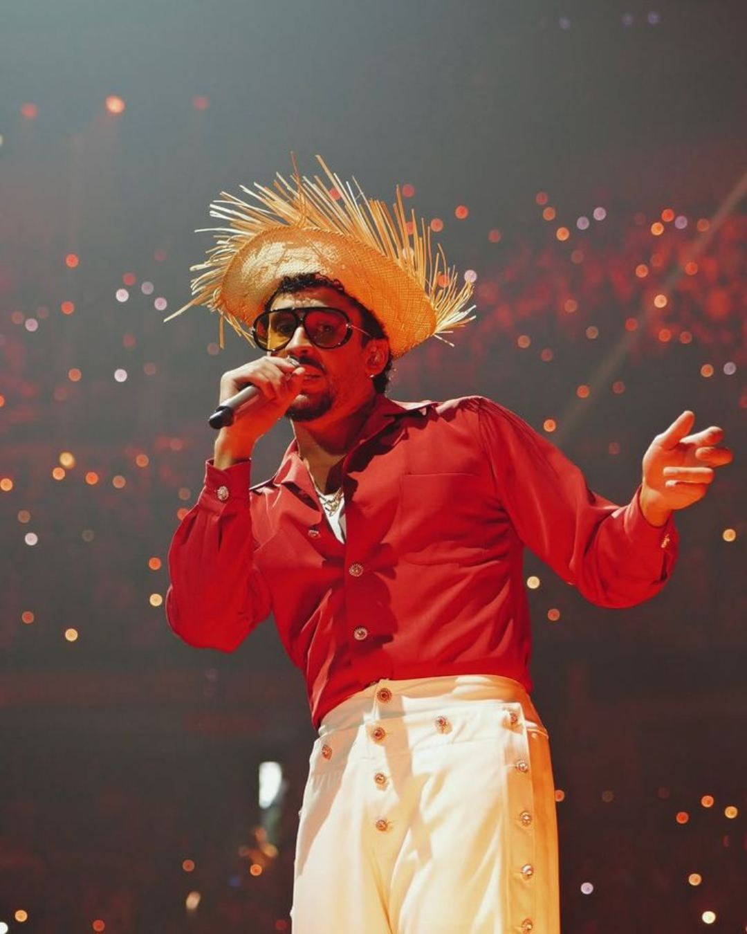 Bad Bunny performing live on stage in a red shirt and straw hat, with bokeh stage lights behind him