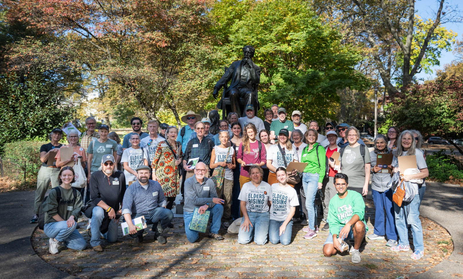 A large group of people stands together in front of a statue in a park. Several people hold clipboards and papers, and trees with autumn foliage surround the group.