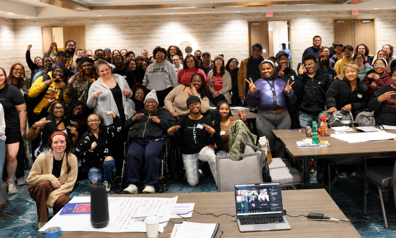A large group of people poses together in a conference room. Tables with notebooks, papers, drinks, and an open laptop are visible in the foreground, and several people smile or make peace signs toward the camera.