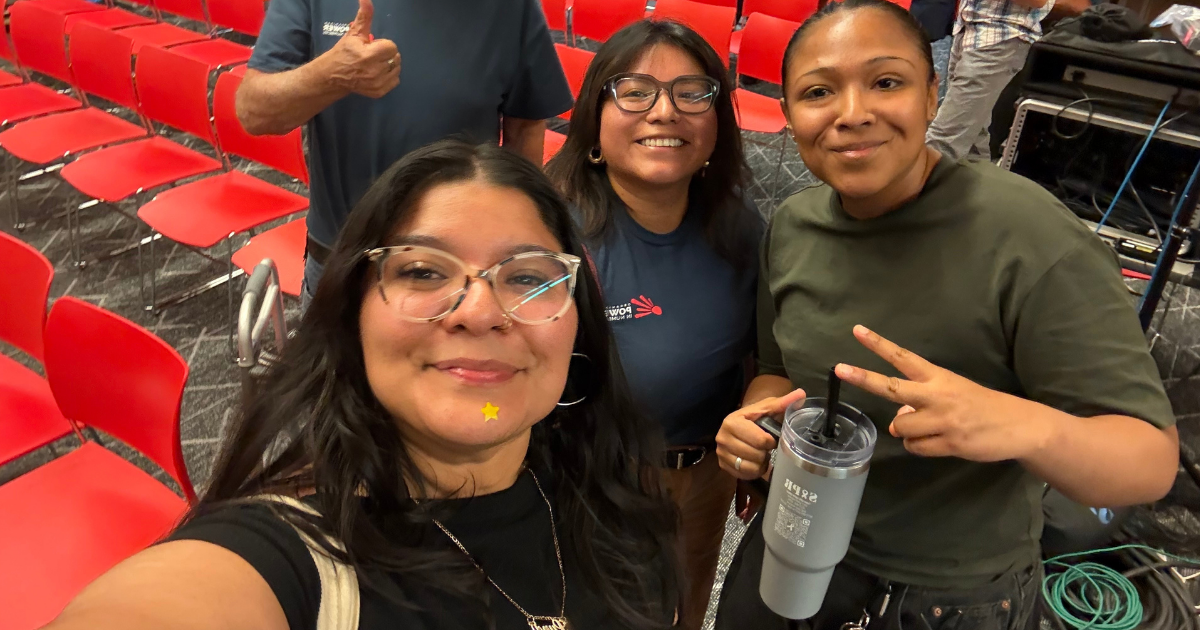 Three Organized Power in Numbers staff and volunteers pose for a selfie after attending a Phoenix City Council meeting. Rows of red chairs are visible behind them, and one person holds up a peace sign.