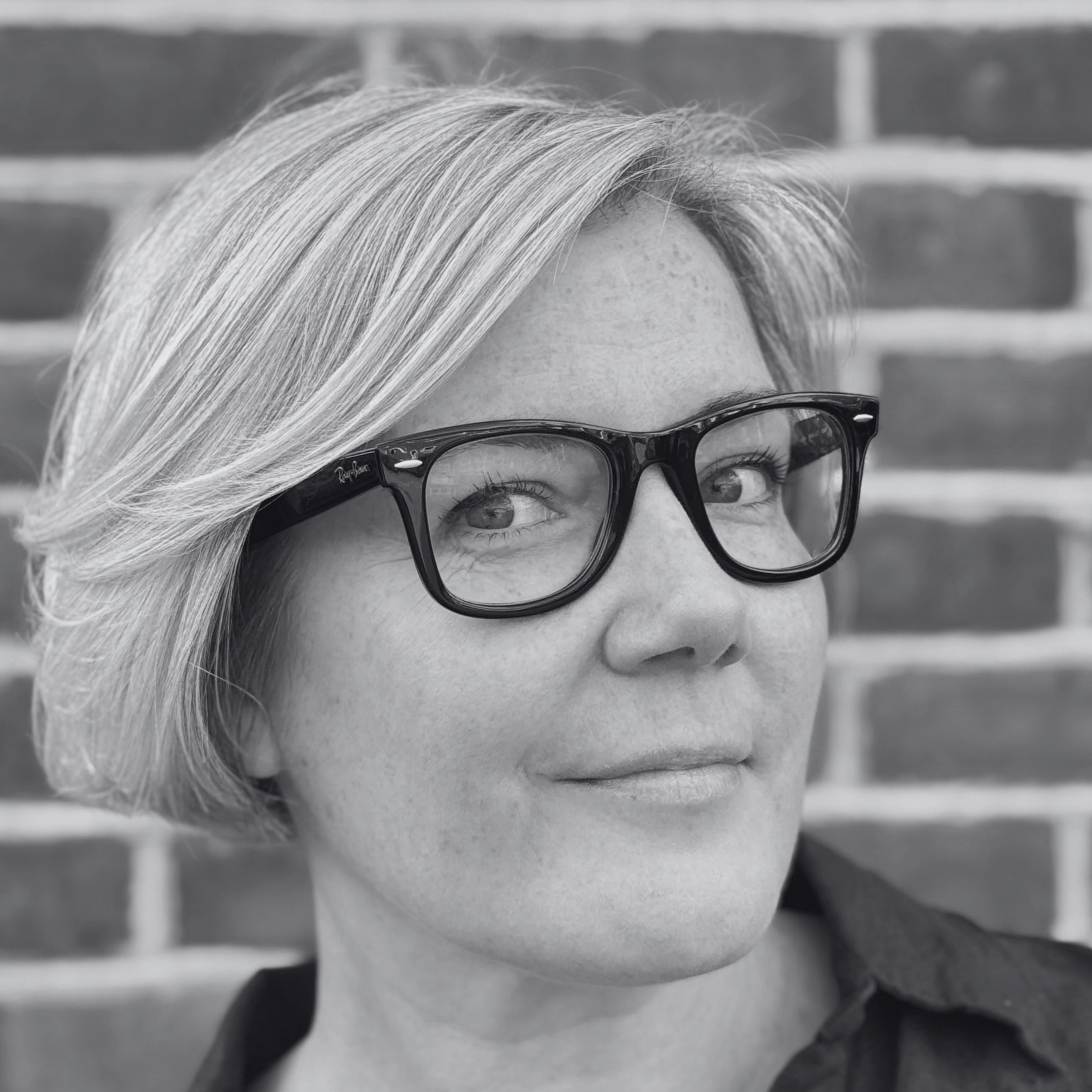 Close-up black and white photo of a person's face wearing glasses, with silver hair and a background of a brick wall.